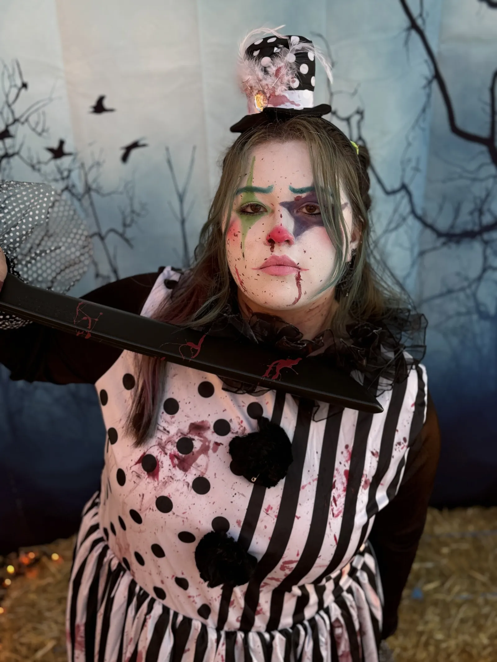 Person dressed as a creepy clown with polka dot and striped outfit, smeared makeup, holding blood-stained sword