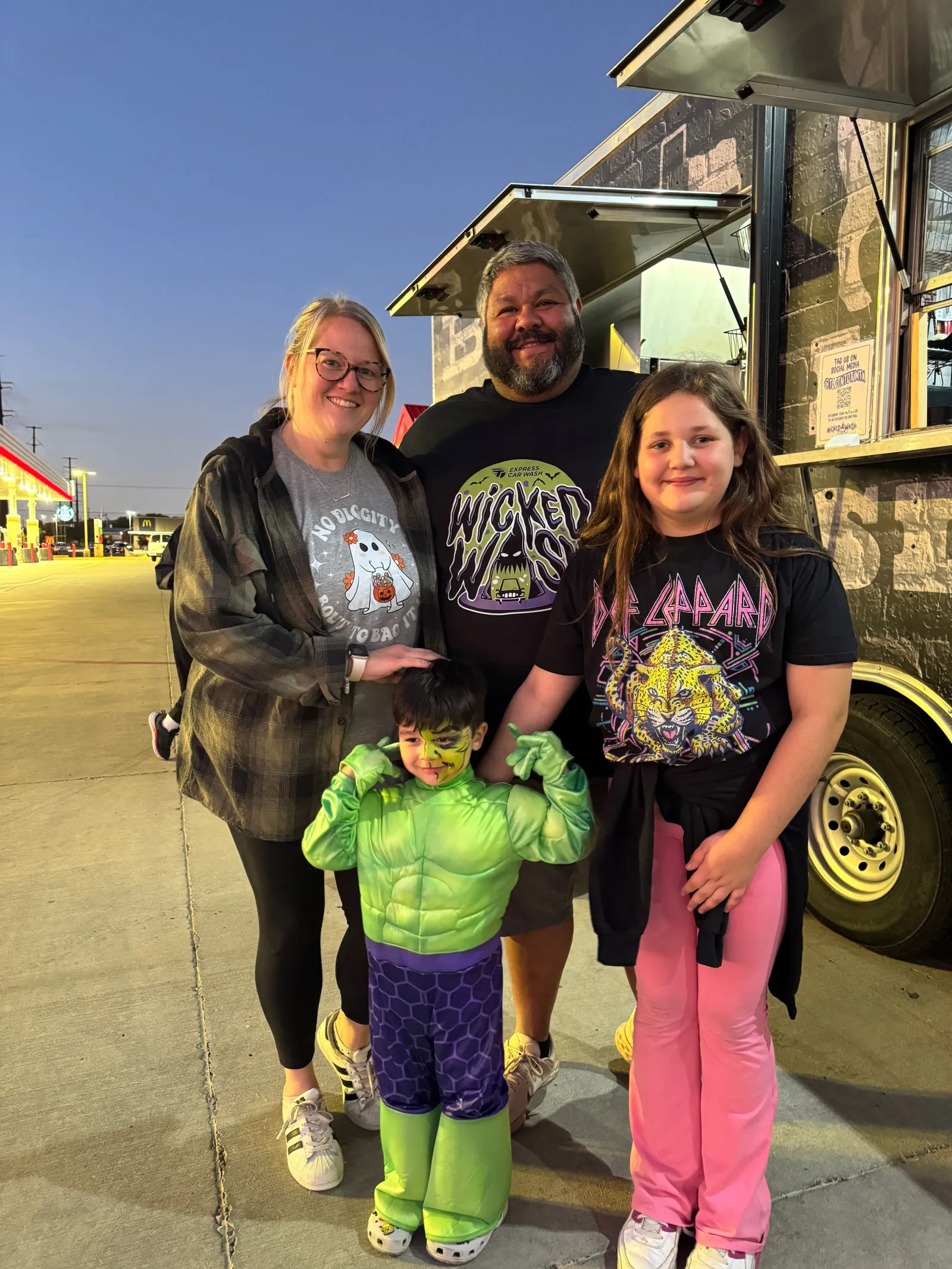 Family of four in casual and costume attire posing near a food truck at dusk outdoors.