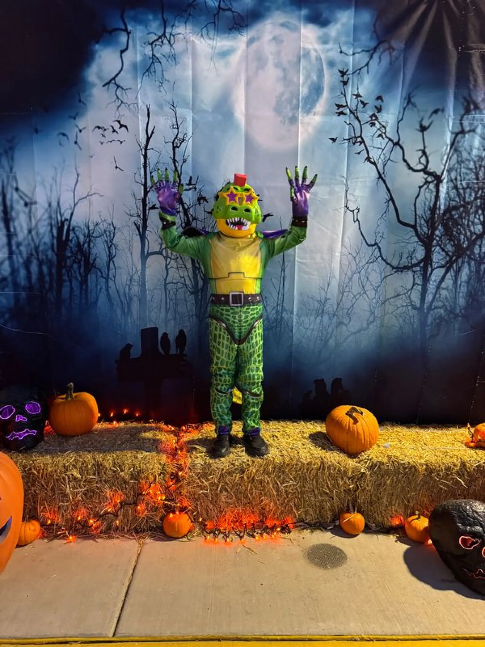 Child in colorful dinosaur costume poses with hands up on hay bale stage decorated with pumpkins and Halloween lights.