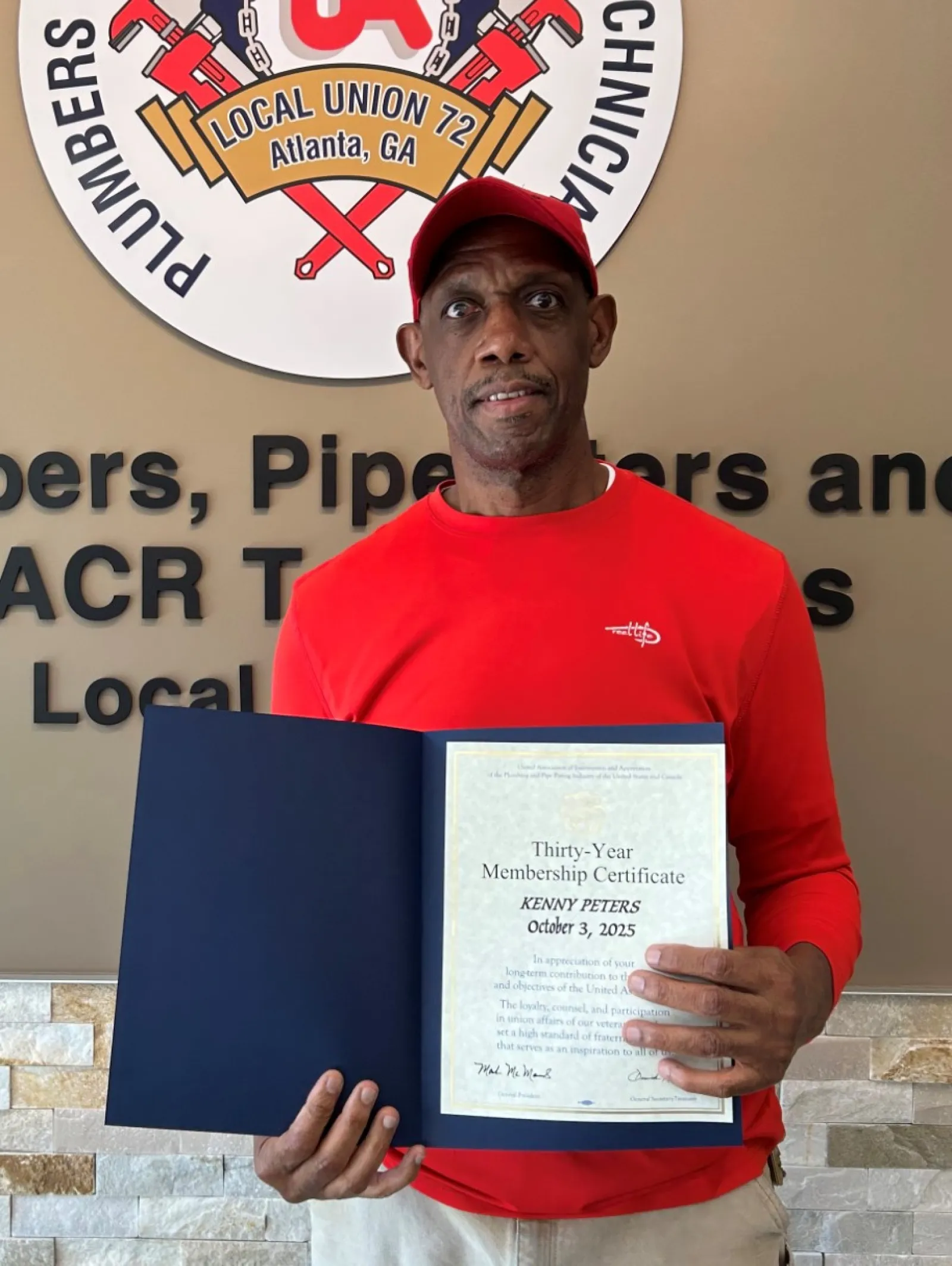 Man in red shirt and cap holding a certification in front of a plumbers and service technicians union sign.