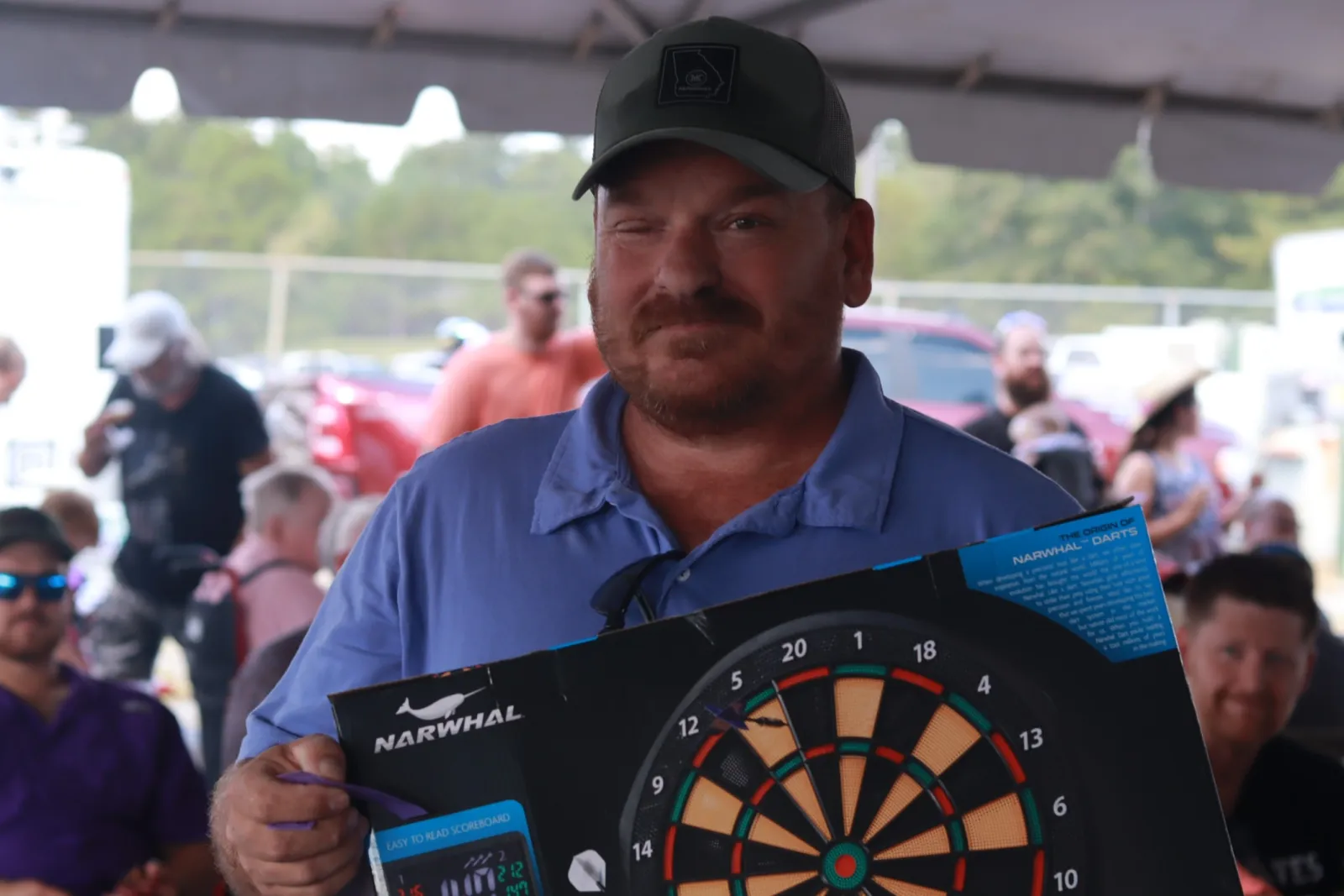 Man in blue shirt and cap holding a Narwhal dartboard with a crowd in background at outdoor event.