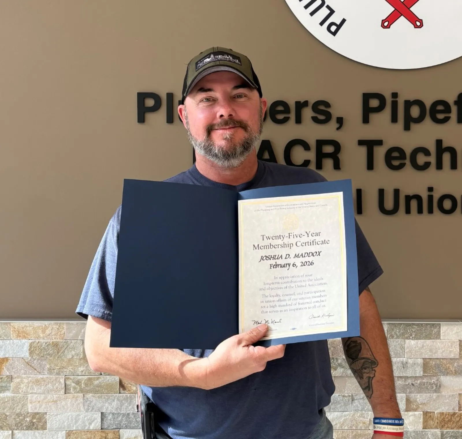 Man in cap holding certificate standing by Plumbers Pipefitters Service Technicians Union logo wall