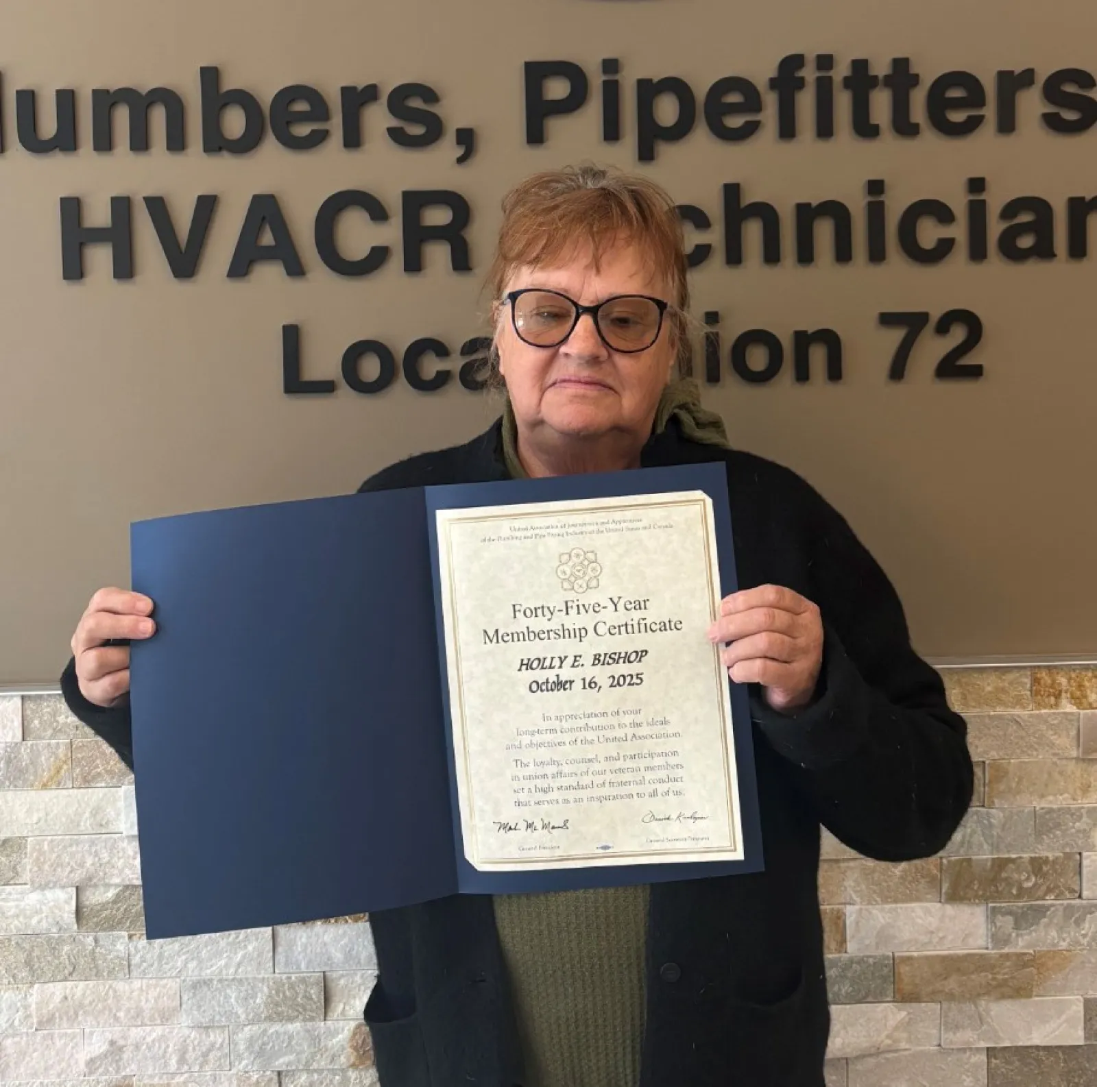 Person holding a certificate standing next to a Plumbers and Pipefitters Union Local 72 sign on a beige wall.