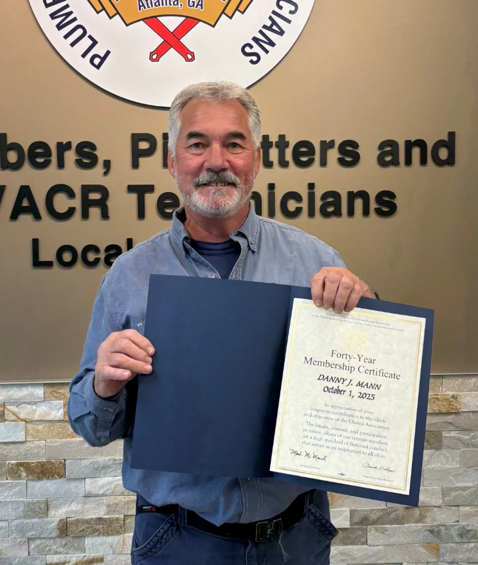Man holding a certificate standing next to Plumbers and Pipefitters Local Union 72 logo on wall