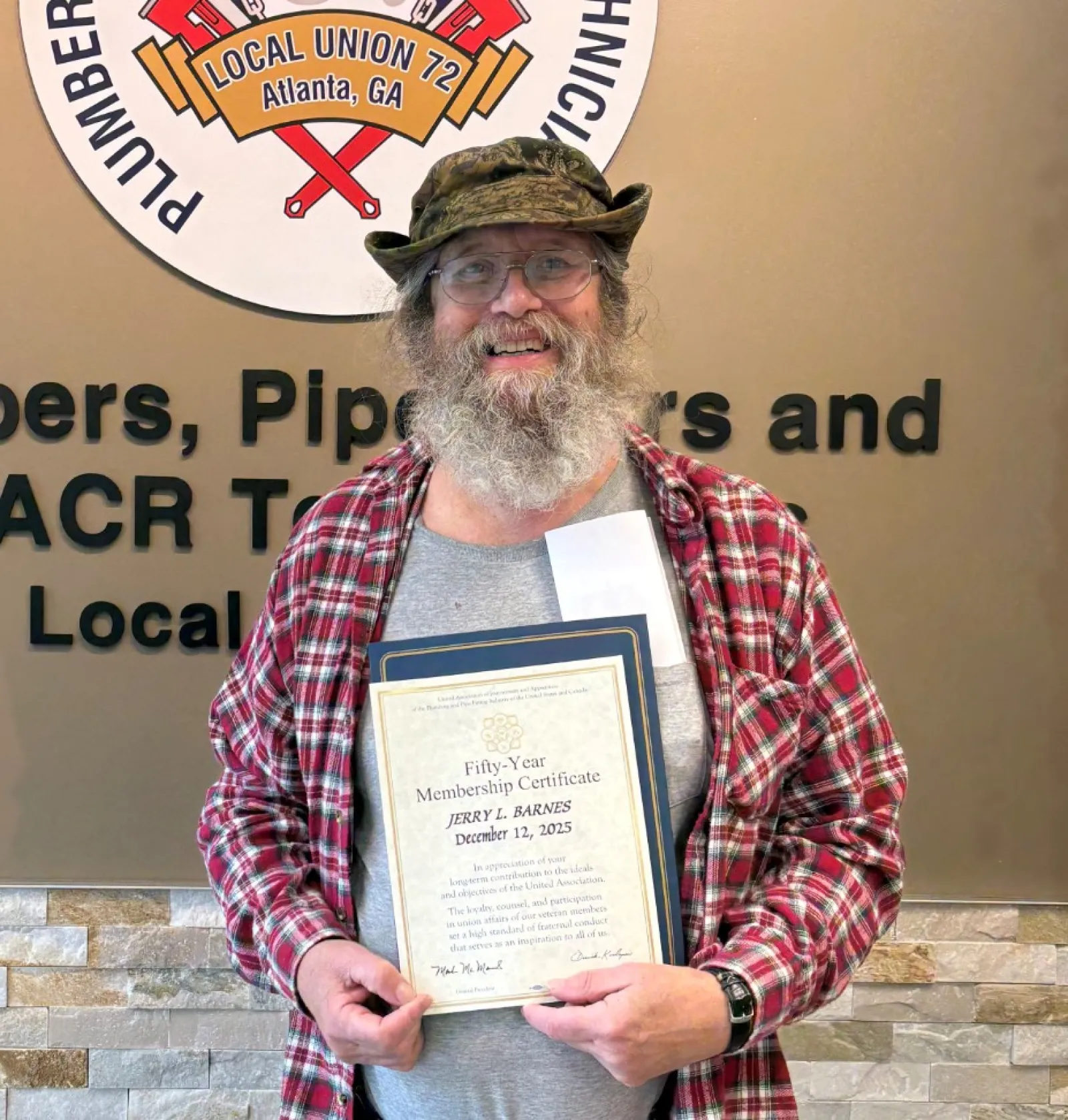 Man in a red plaid shirt holding a certificate standing next to a Plumbers service union sign on a wall.