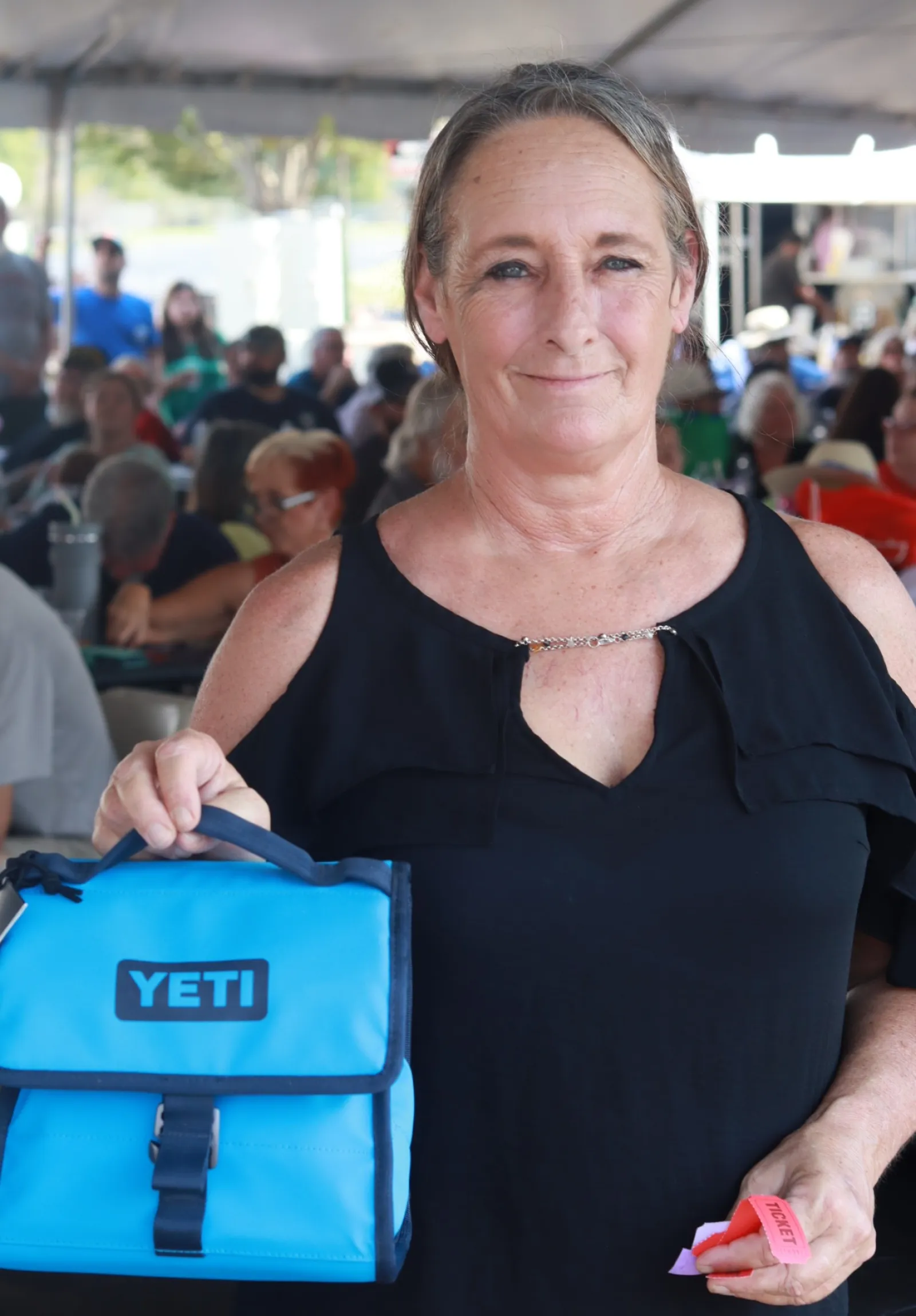 Woman holding a blue YETI cooler bag and event tickets at an outdoor gathering under a tent.
