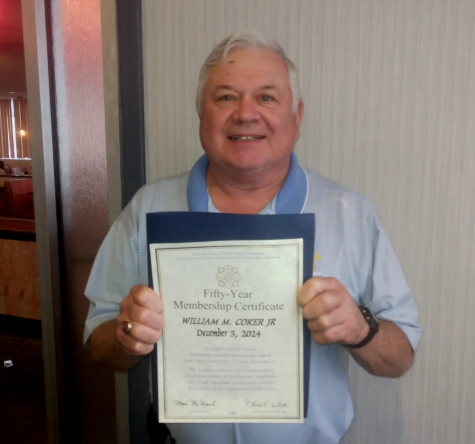 Man holding a fifty-year membership certificate with a smile, standing indoors against a neutral wall.