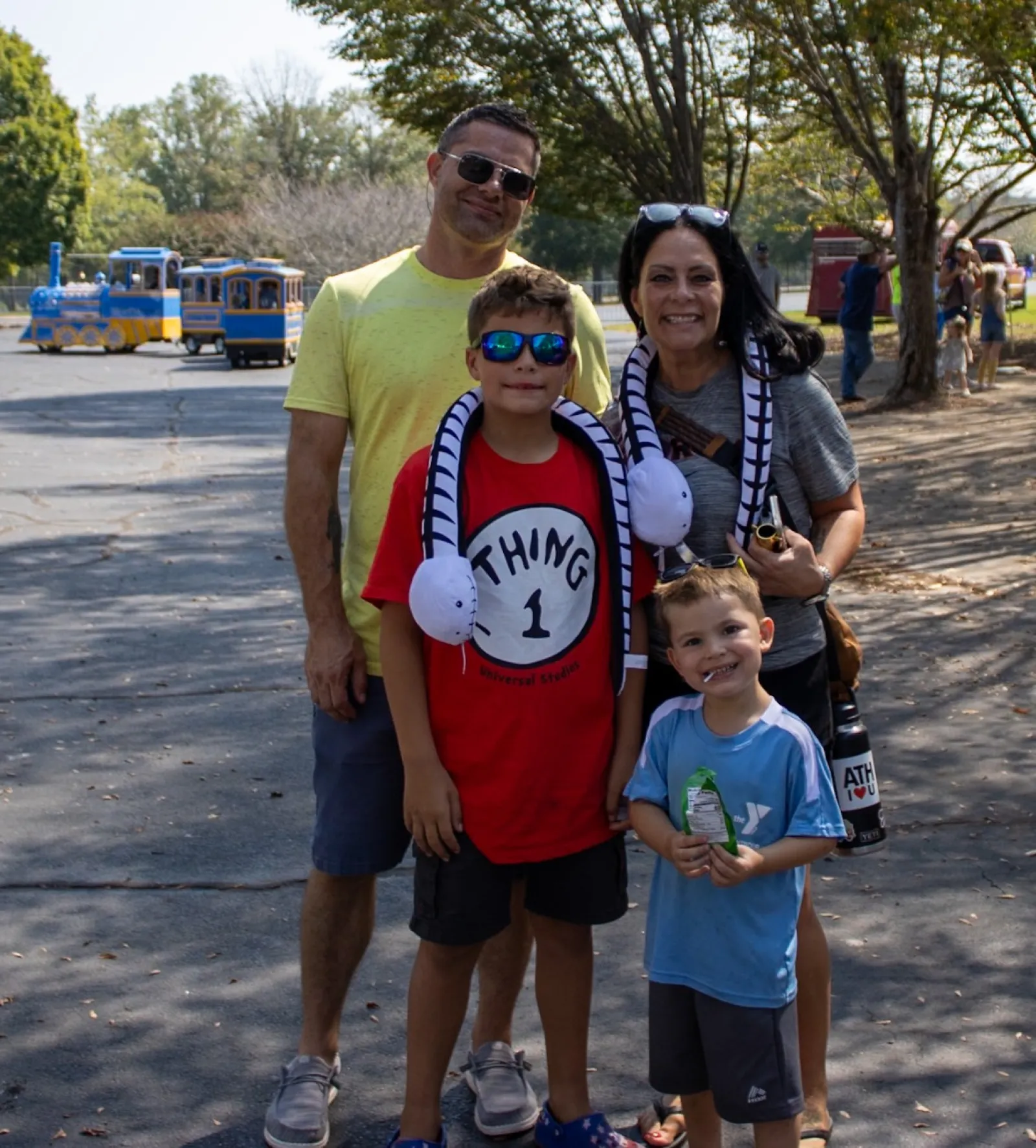 Smiling family of four posing outdoors on a sunny day with trees and a small train in the background.