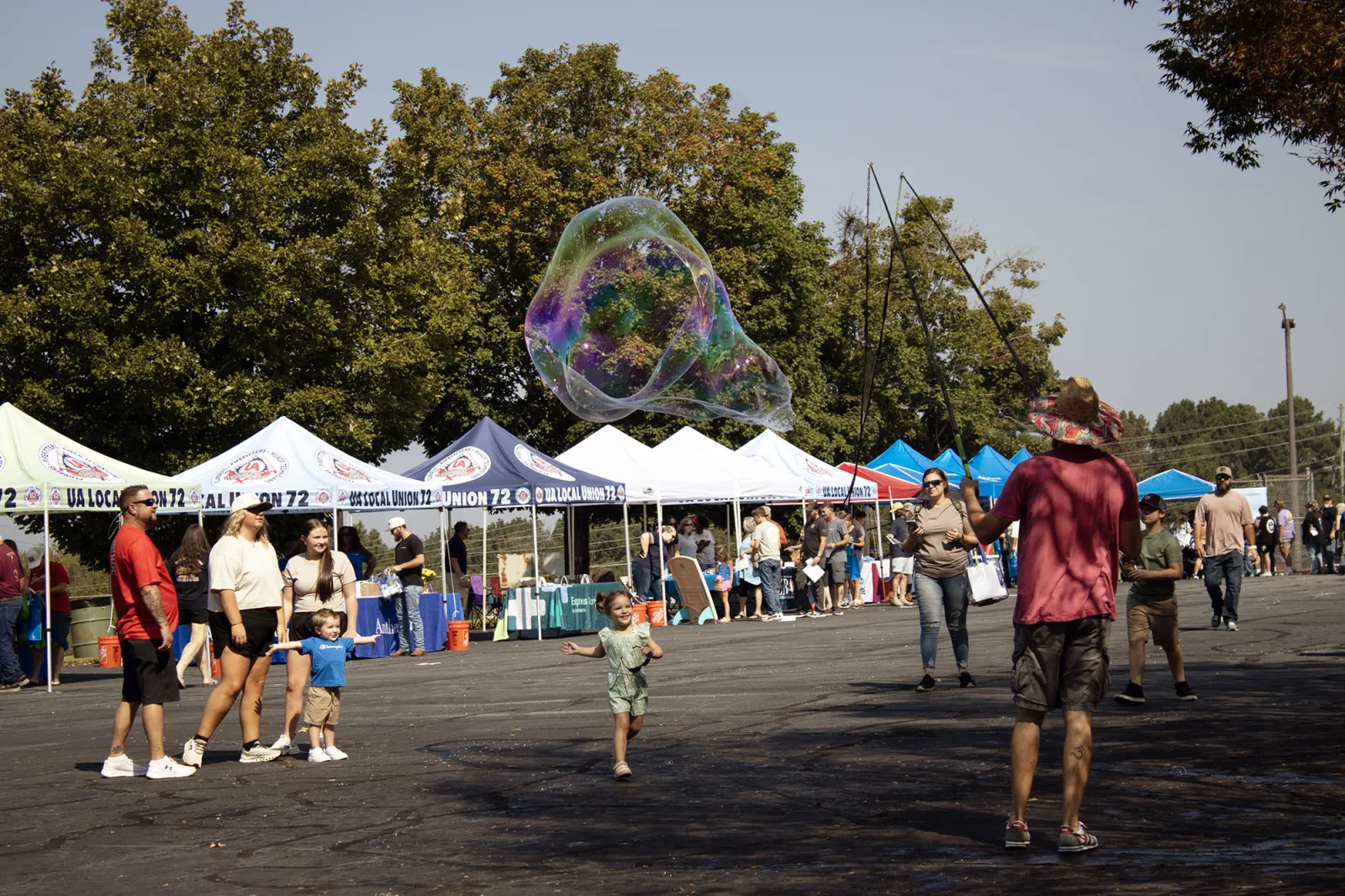 Children and families enjoy a sunny outdoor market with large soap bubbles and vendor tents in the background.