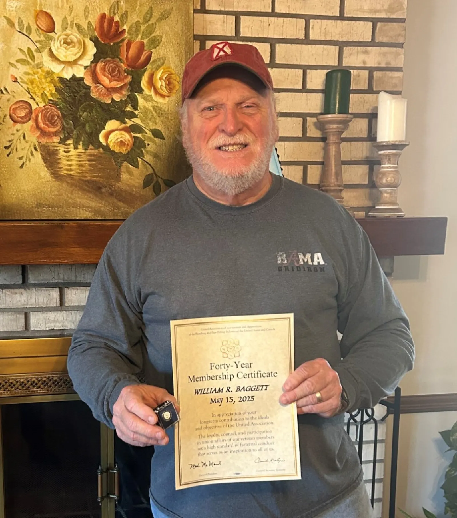 Smiling man holding a certificate standing by a brick fireplace with floral painting and plants.