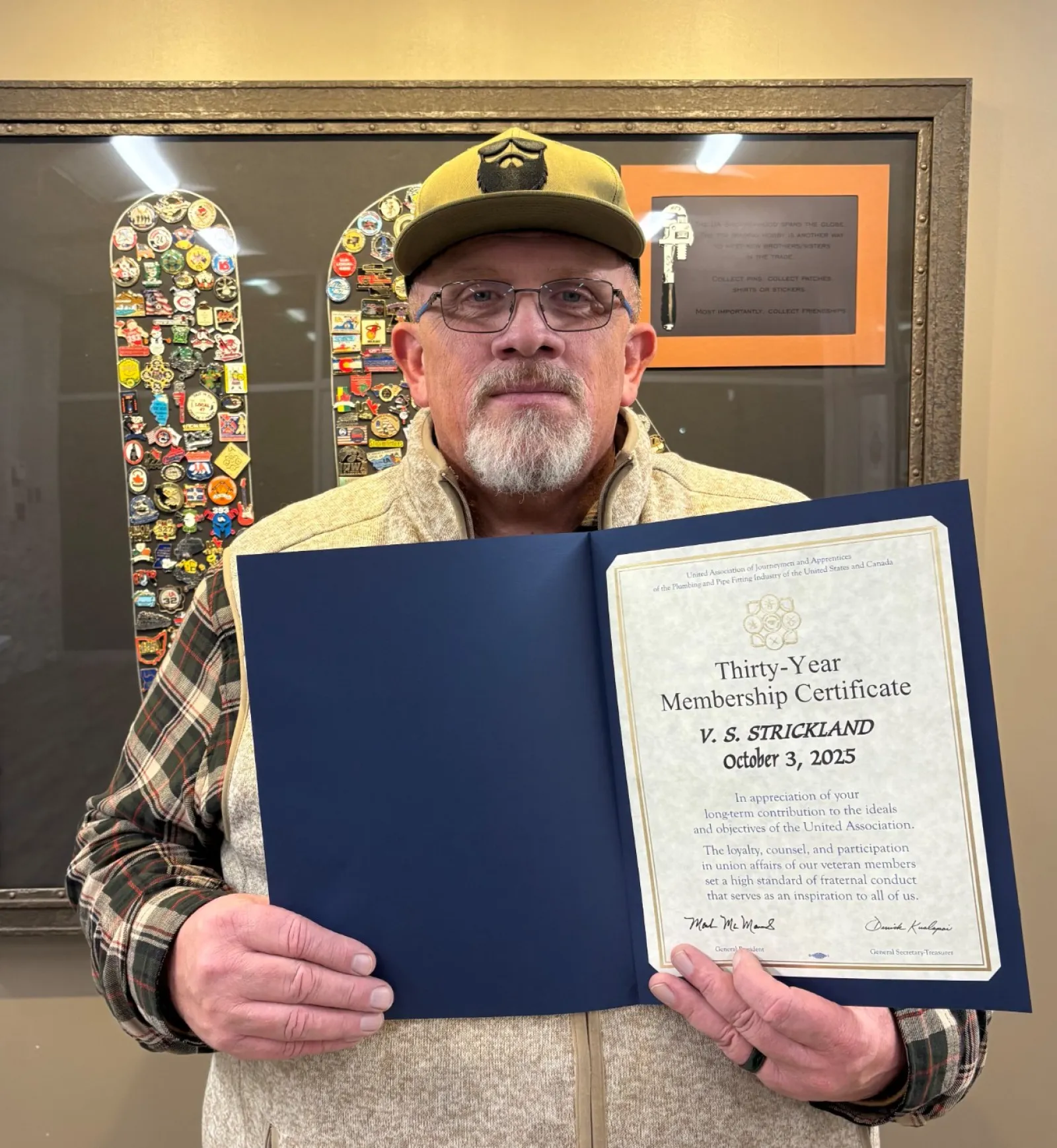 Man with beard and glasses holding a thirty-year membership certificate in front of a framed pin collection.