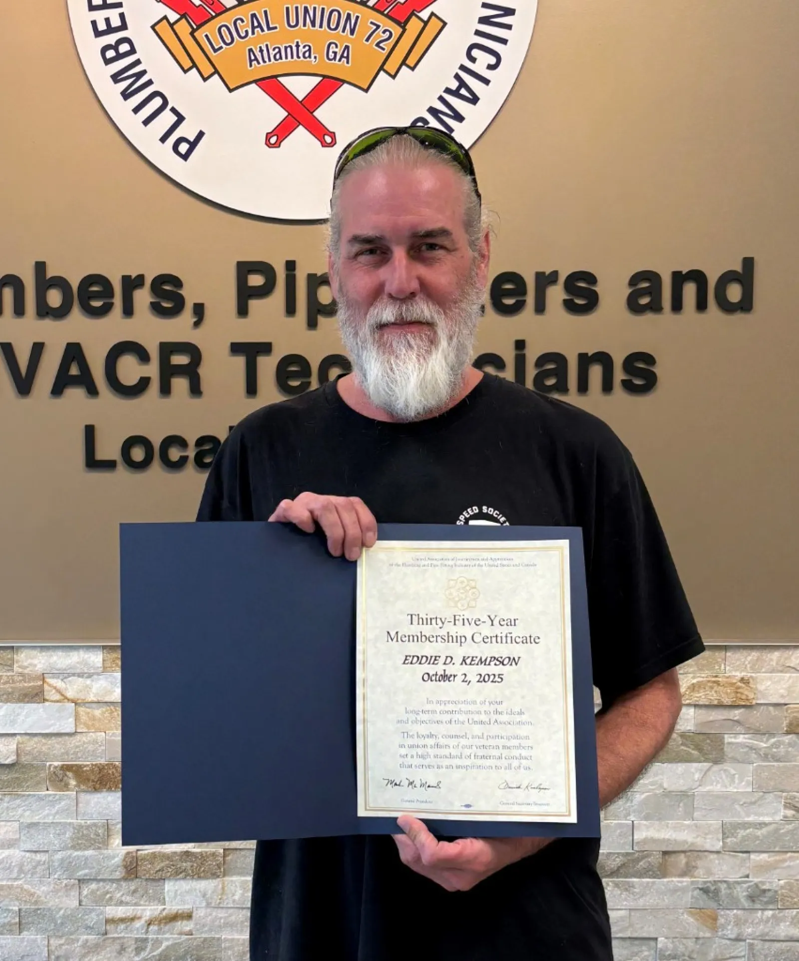 Man with gray beard holding a certificate standing in front of a Plumbers and Pipefitters union sign and wall