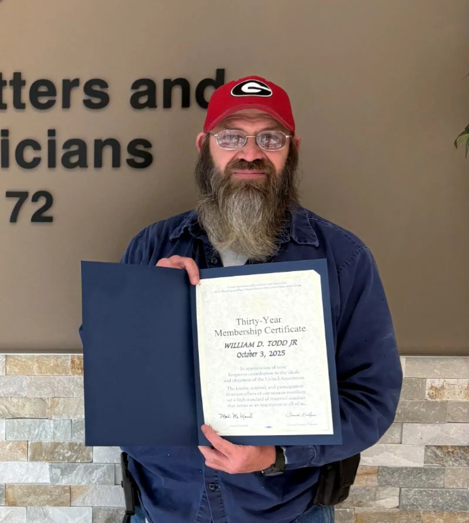 Bearded man in red hat holding certificate standing next to Plumbers, Pipefitters & HVACR Technicians Local Union 72 logo.