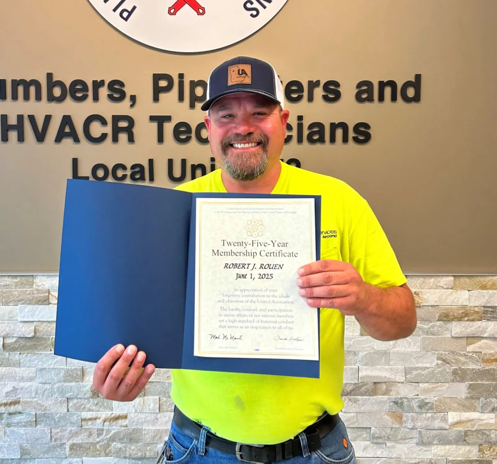 Smiling man in yellow shirt holding a certificate standing beside a Plumbers and Pipefitters union sign on wall.