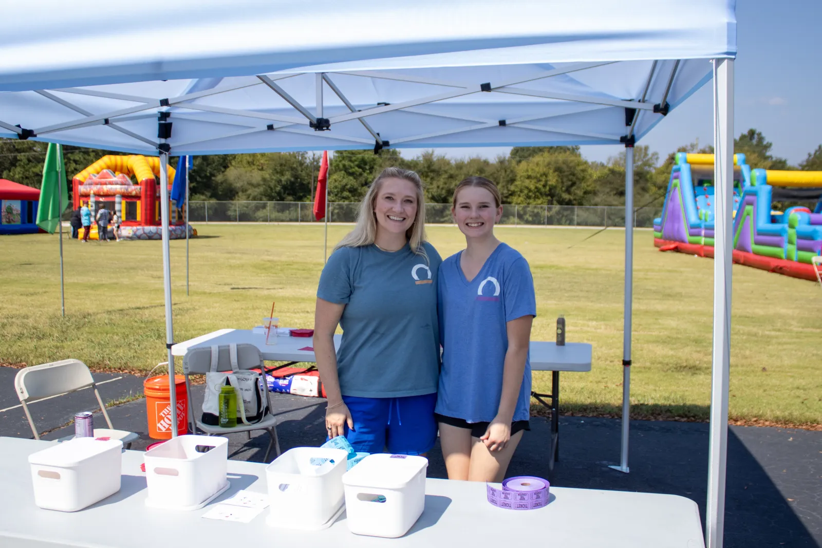 Two women smiling under a white canopy tent at an outdoor event with inflatable attractions in the background