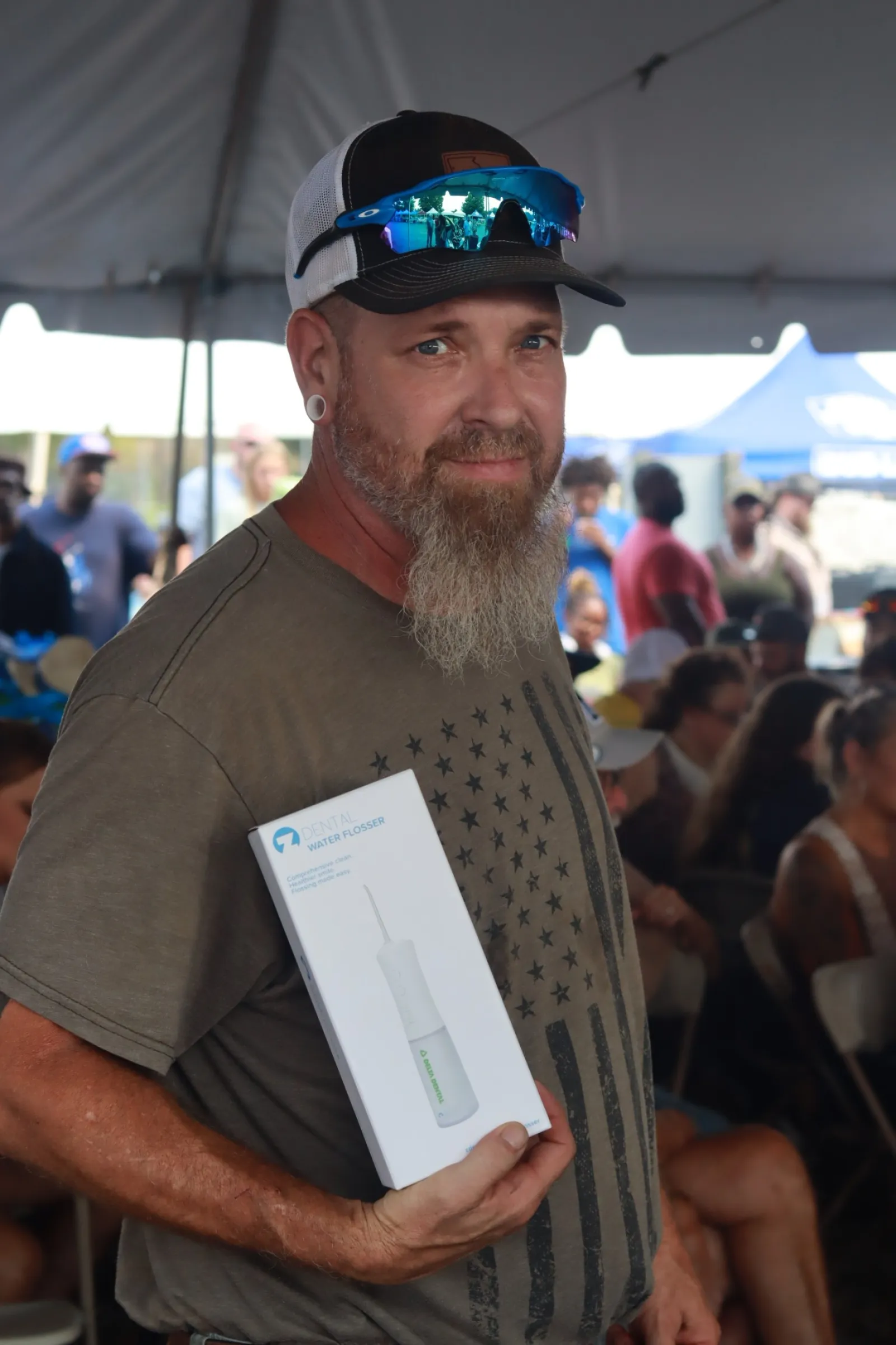 Man with a beard and sunglasses holding a white product box in a crowded event tent