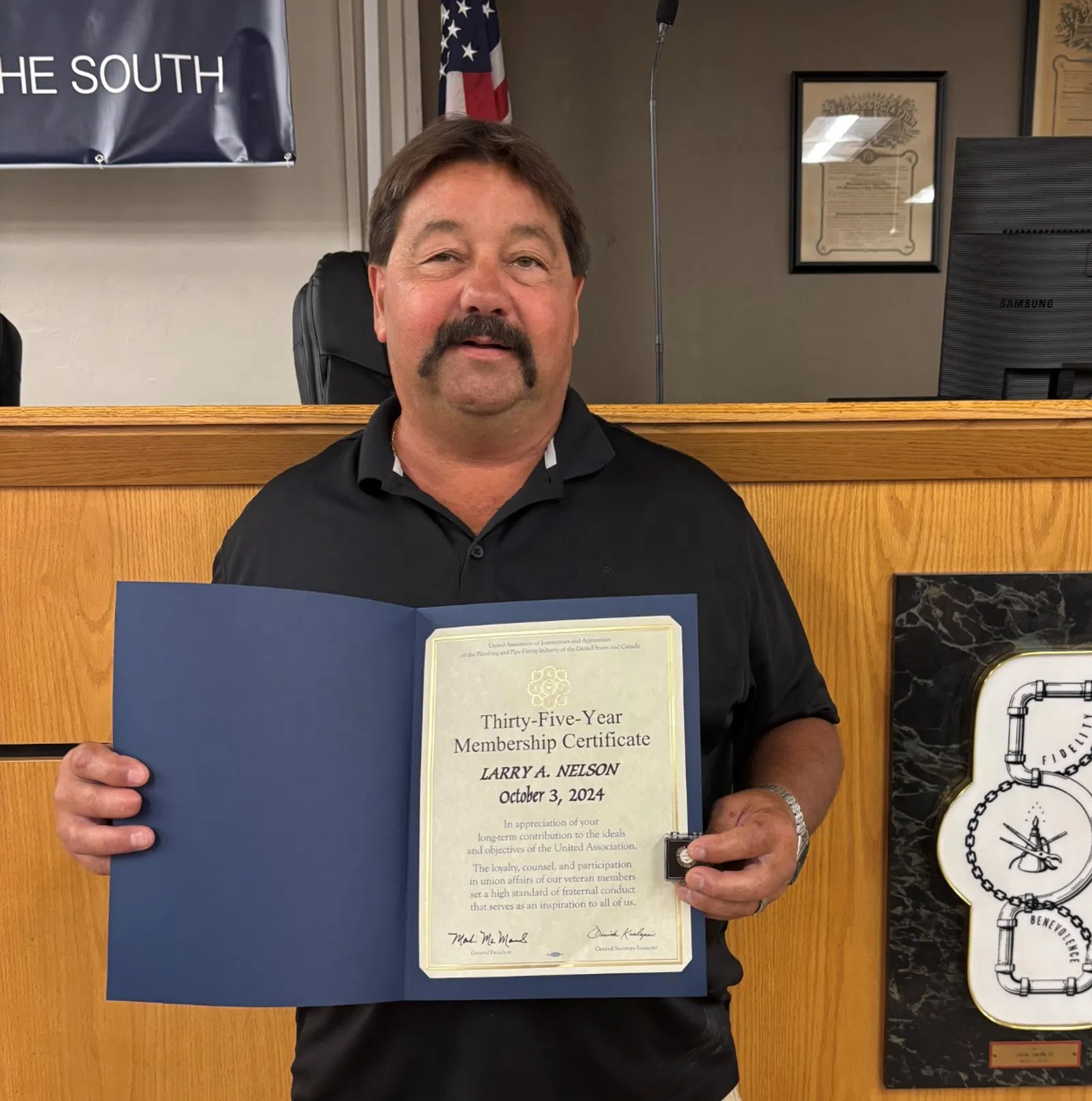 Man in black shirt and shorts holding a certificate in a formal office with wooden paneling and an American flag.