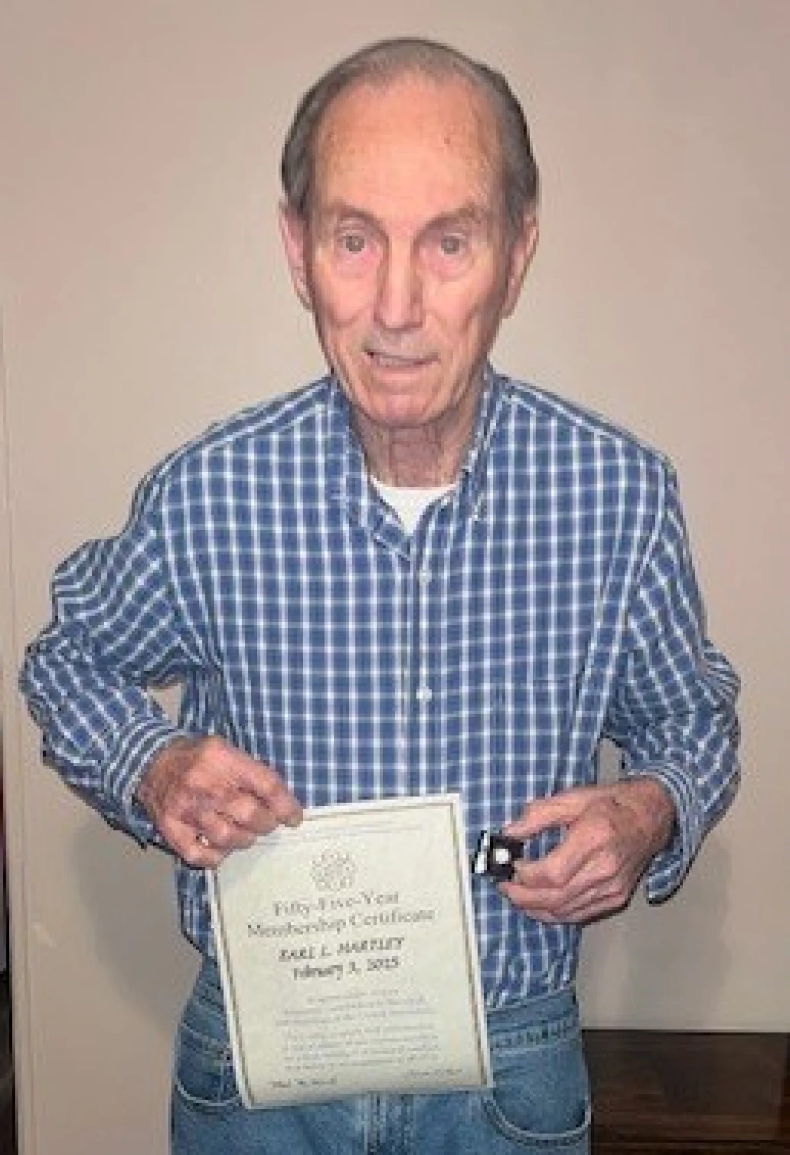 Elderly man in blue plaid shirt holding a certificate and a small black box, standing indoors on wood floor.