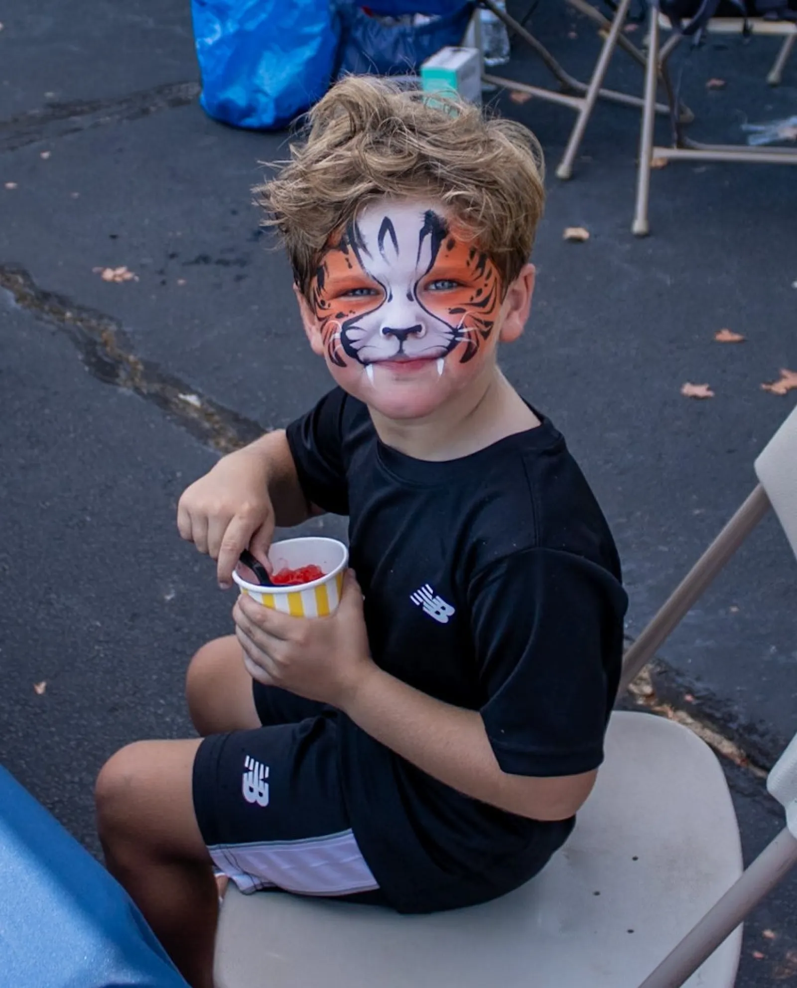 Young boy with tiger face paint sitting on a chair holding a cup of snacks outdoors at a community event.