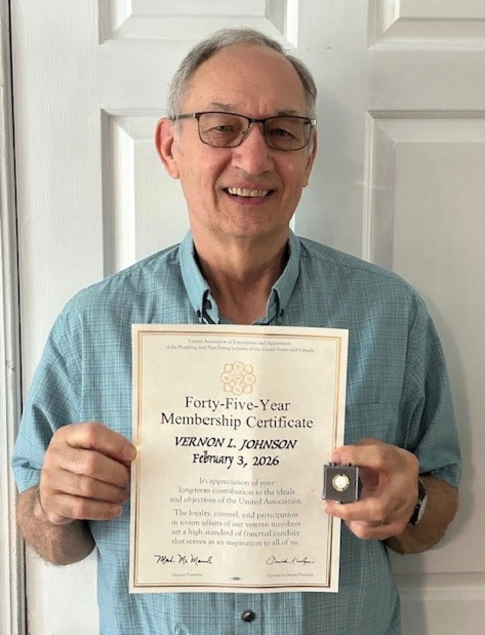 Man wearing glasses and blue shirt holding a forty-five year membership certificate and a pin, smiling at the camera.