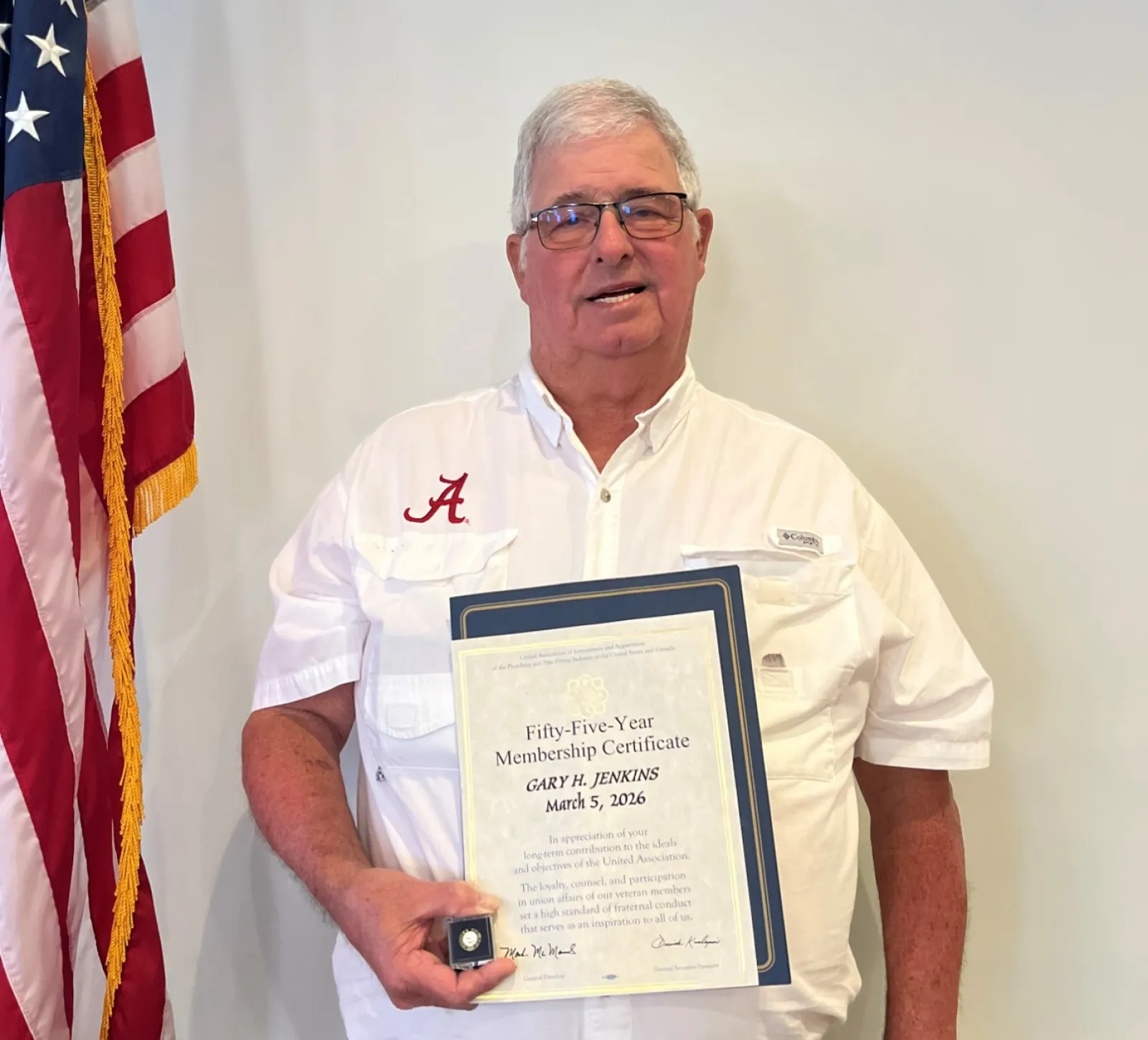 Man in white shirt holding a certificate and medal standing next to an American flag indoors.