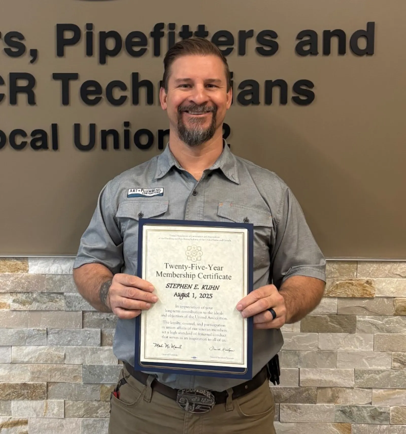 Man in uniform holding certificate standing by Plumbers and Pipefitters Local Union 72 sign on wall.