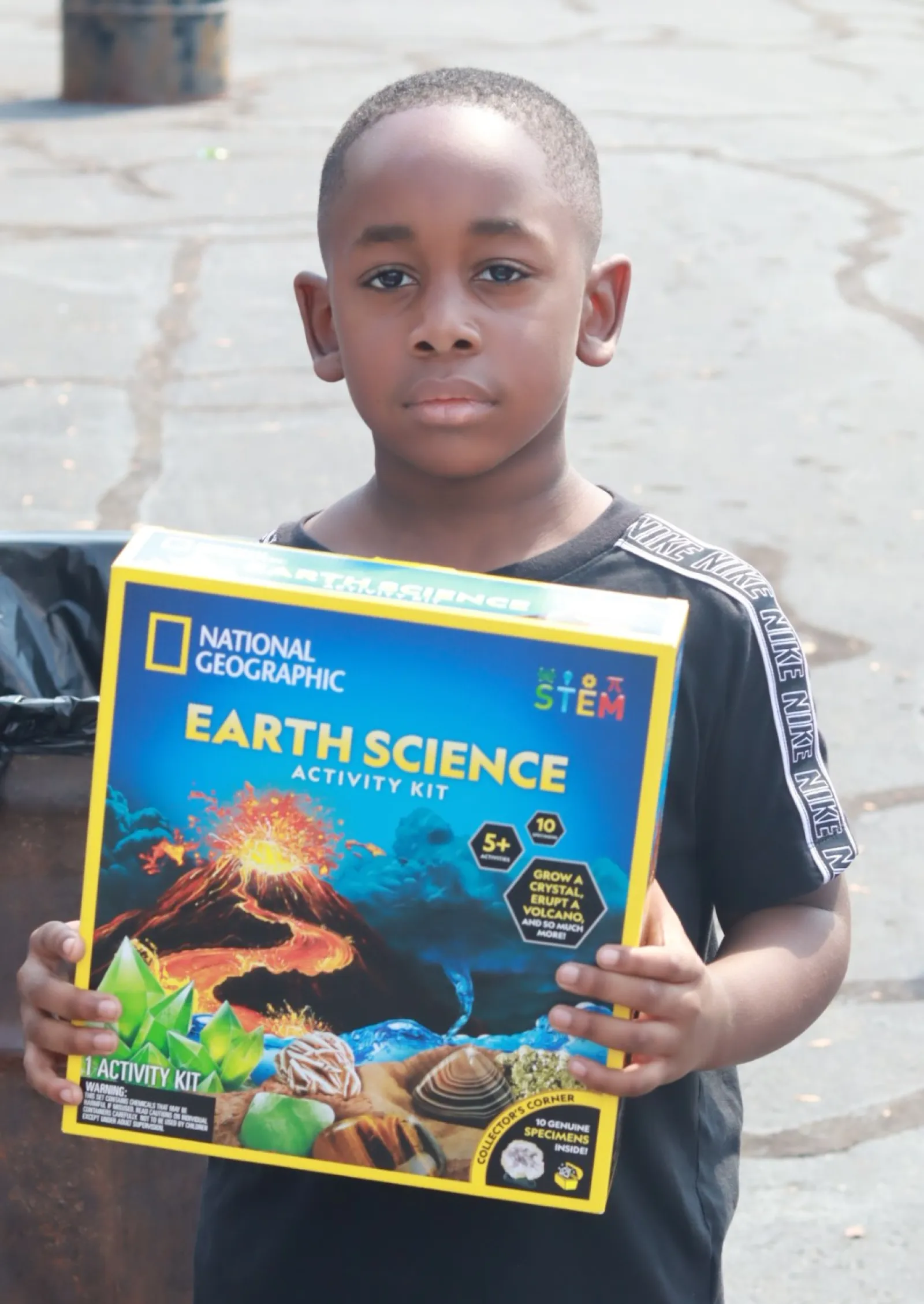 Young boy holding a National Geographic Earth Science activity kit standing outdoors on a paved area.