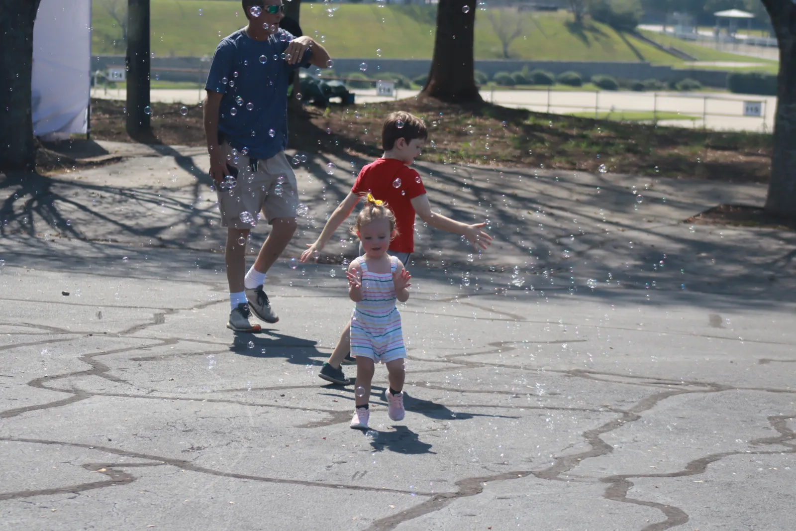 Young children and an adult playing with bubbles outdoors on a sunny day in a paved area with trees.