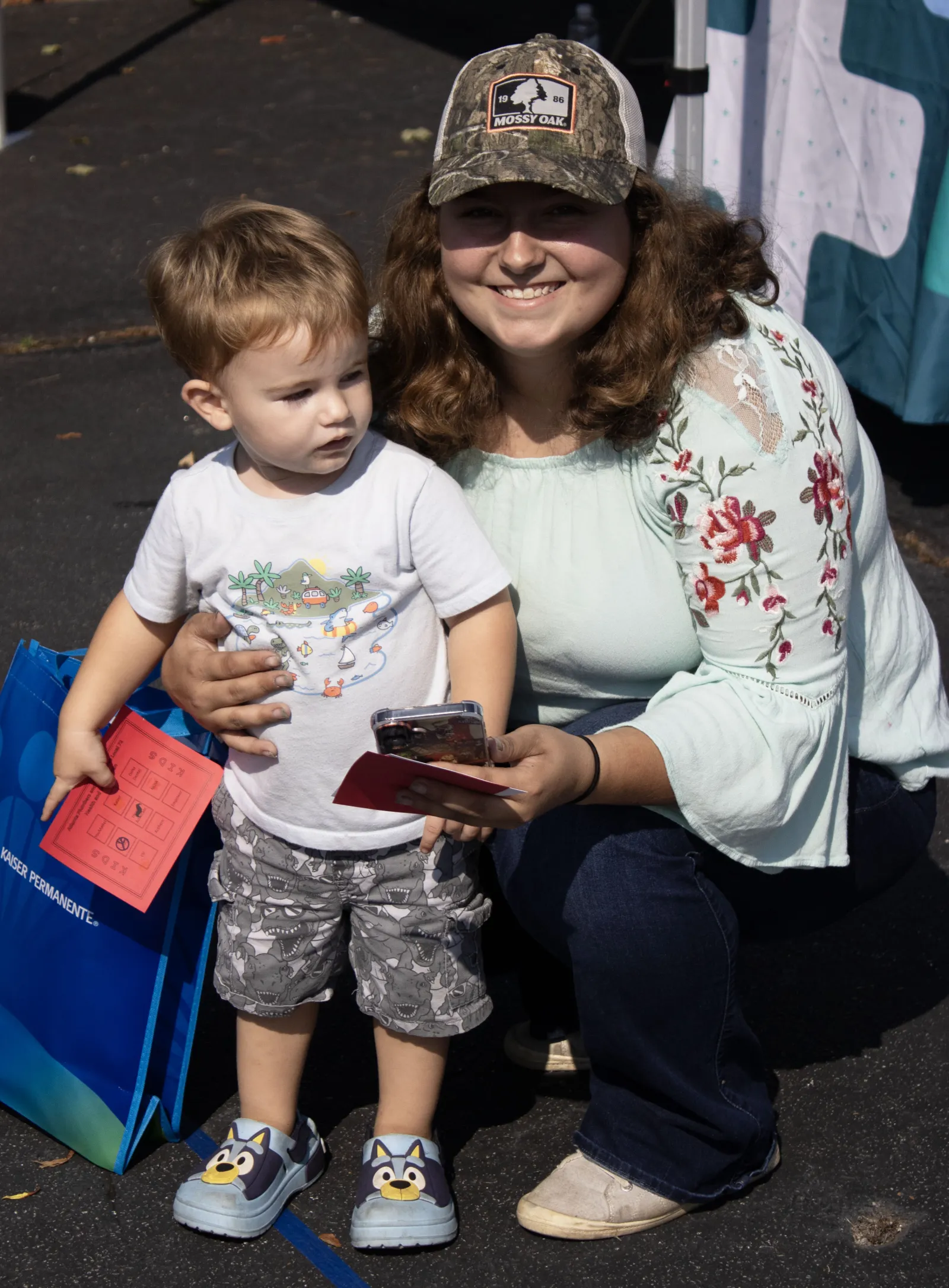 Smiling woman with floral blouse and camo hat kneels next to toddler holding red paper and phone outside on pavement.