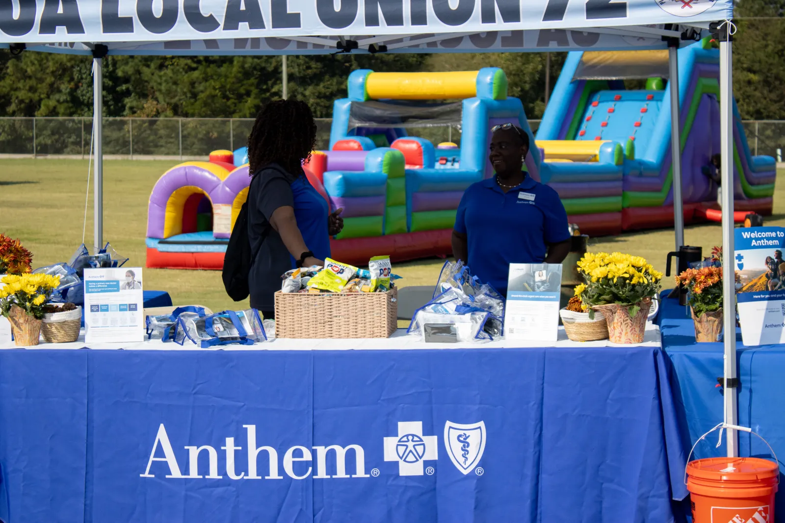 Two women at an Anthem booth with snacks and flowers in front of a blue inflatable play area outdoors