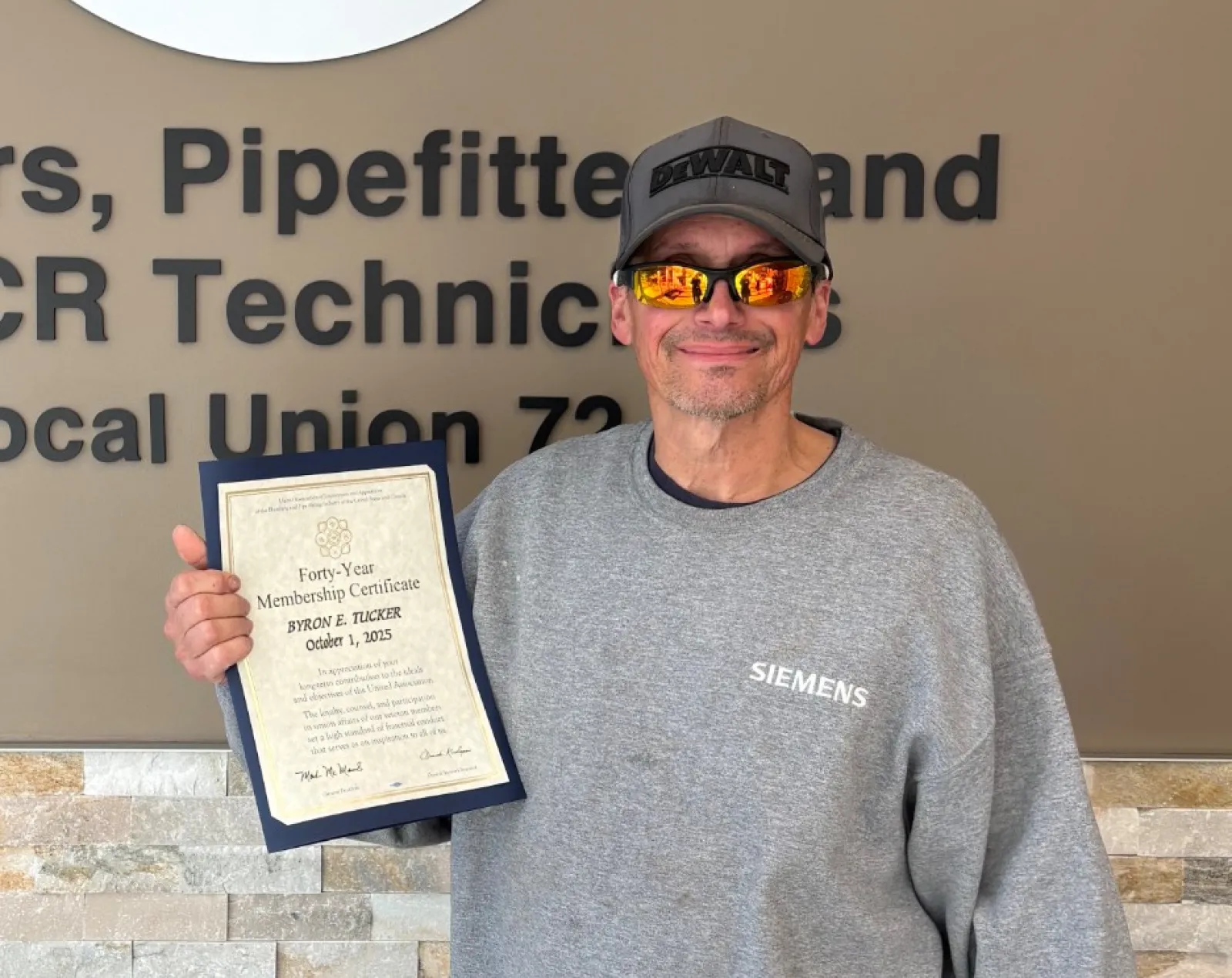 Man wearing sunglasses and a Siemens sweatshirt holding a certificate next to a Plumbing and HVAC union sign.
