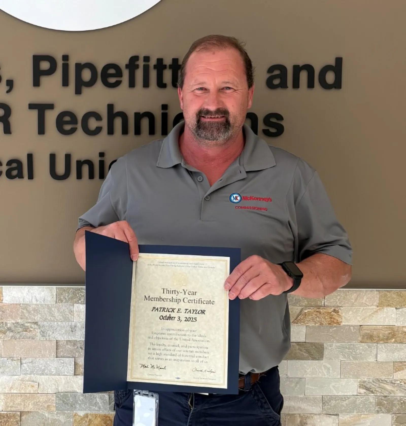 Man proudly holding certificate standing by Plumbers, Pipefitters & Service Technicians union sign on wall