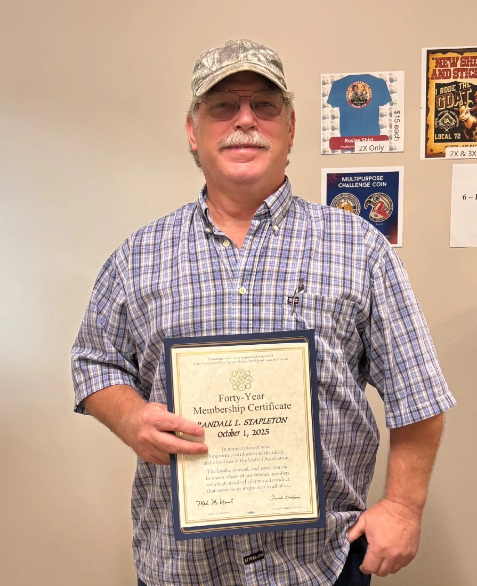 Man in plaid shirt and cap holding a framed certificate standing in a room with posters on the wall.