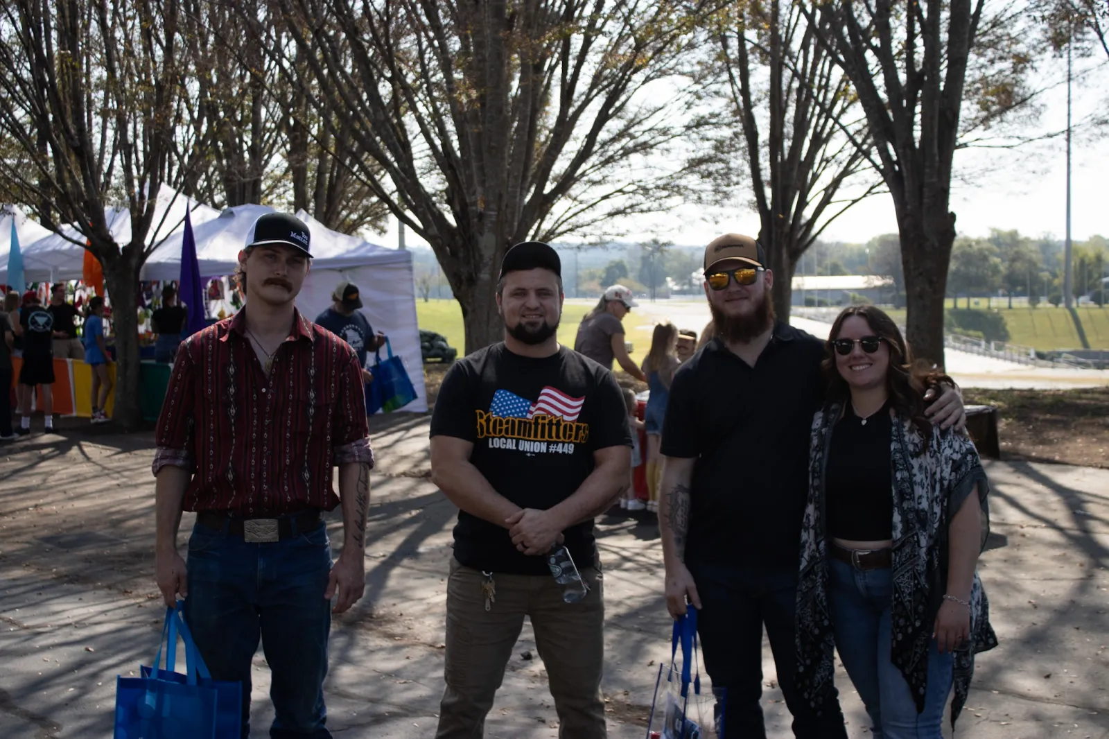 Four friends standing outdoors at a sunny event with tents and trees in the background, holding blue bags.