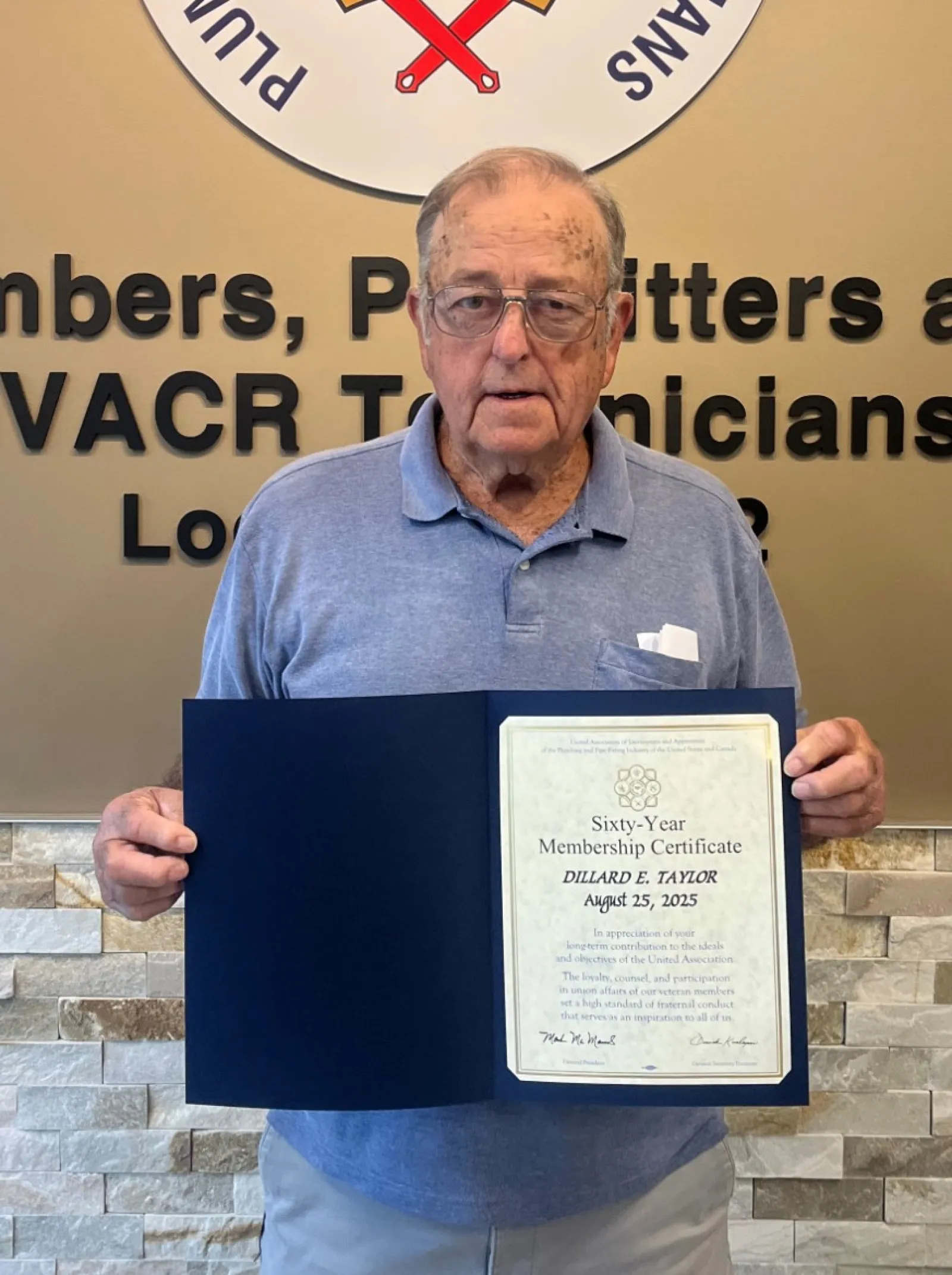 Elderly man holding a certificate standing next to a Plumbers and Pipefitters union emblem on a beige wall.