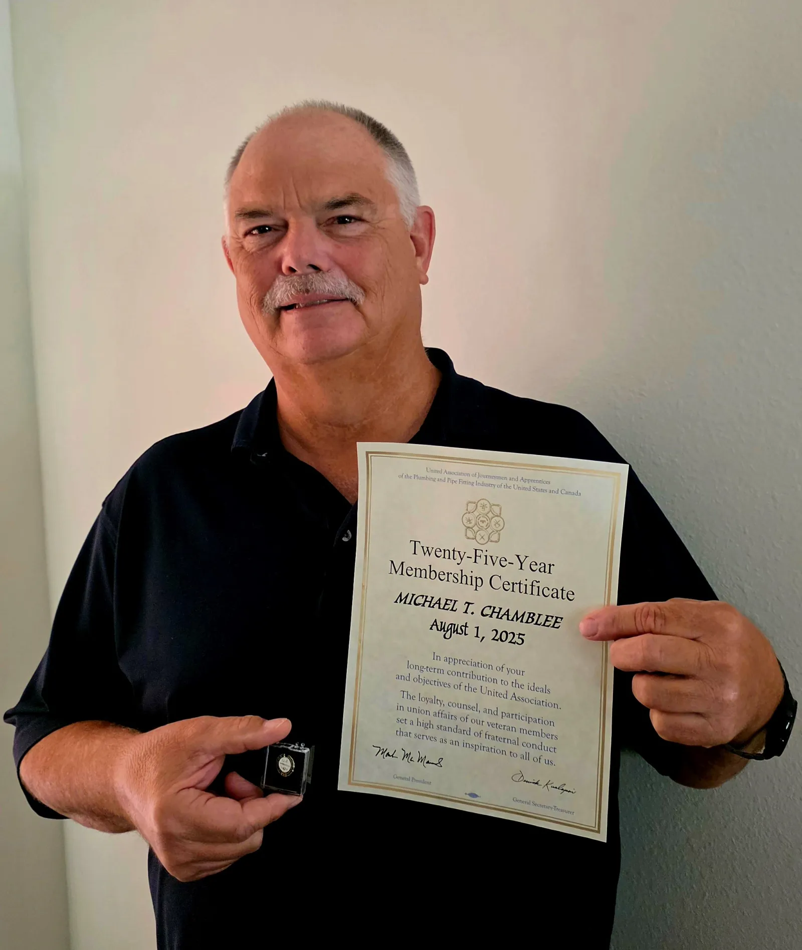 Man holding a Twenty-Five-Year Membership Certificate and a lapel pin in front of a plain wall.