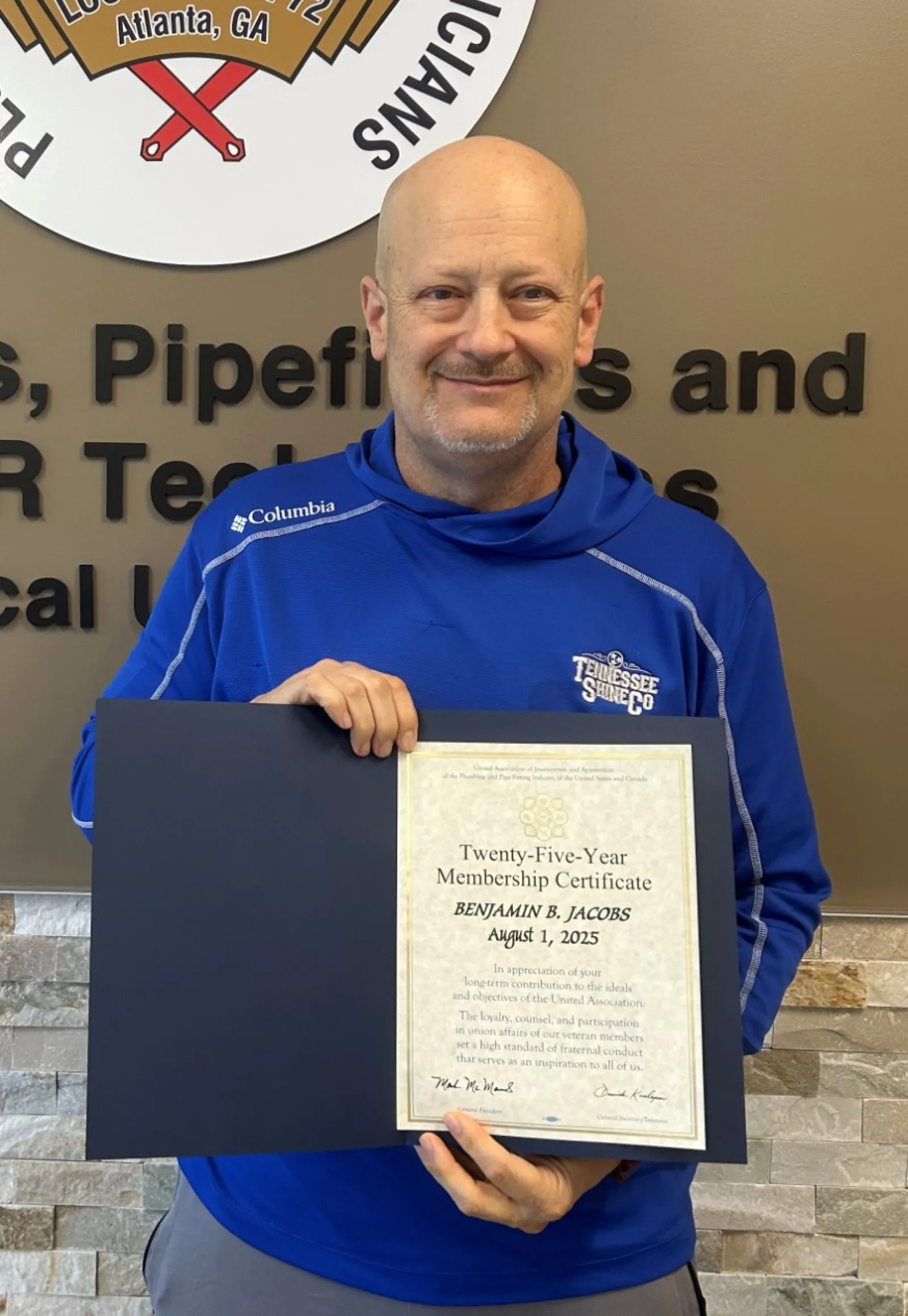Man in blue jacket holding certificate in front of Plumbers and Pipefitters Union Local 72 logo and wall text