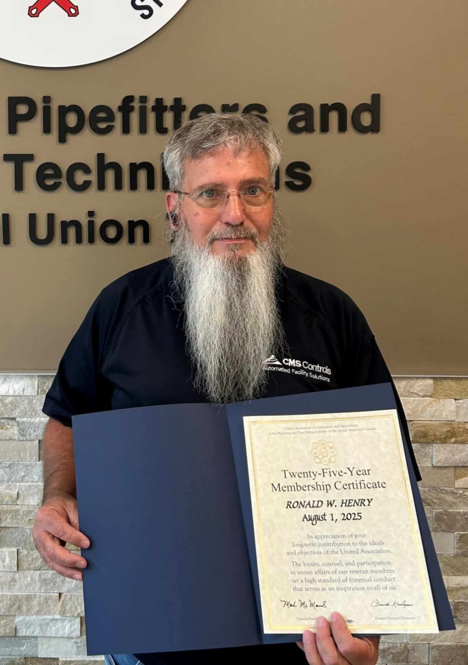 Man with long beard holding a certificate in front of Plumbers, Pipefitters & HVACR Technicians Local Union 72 sign.