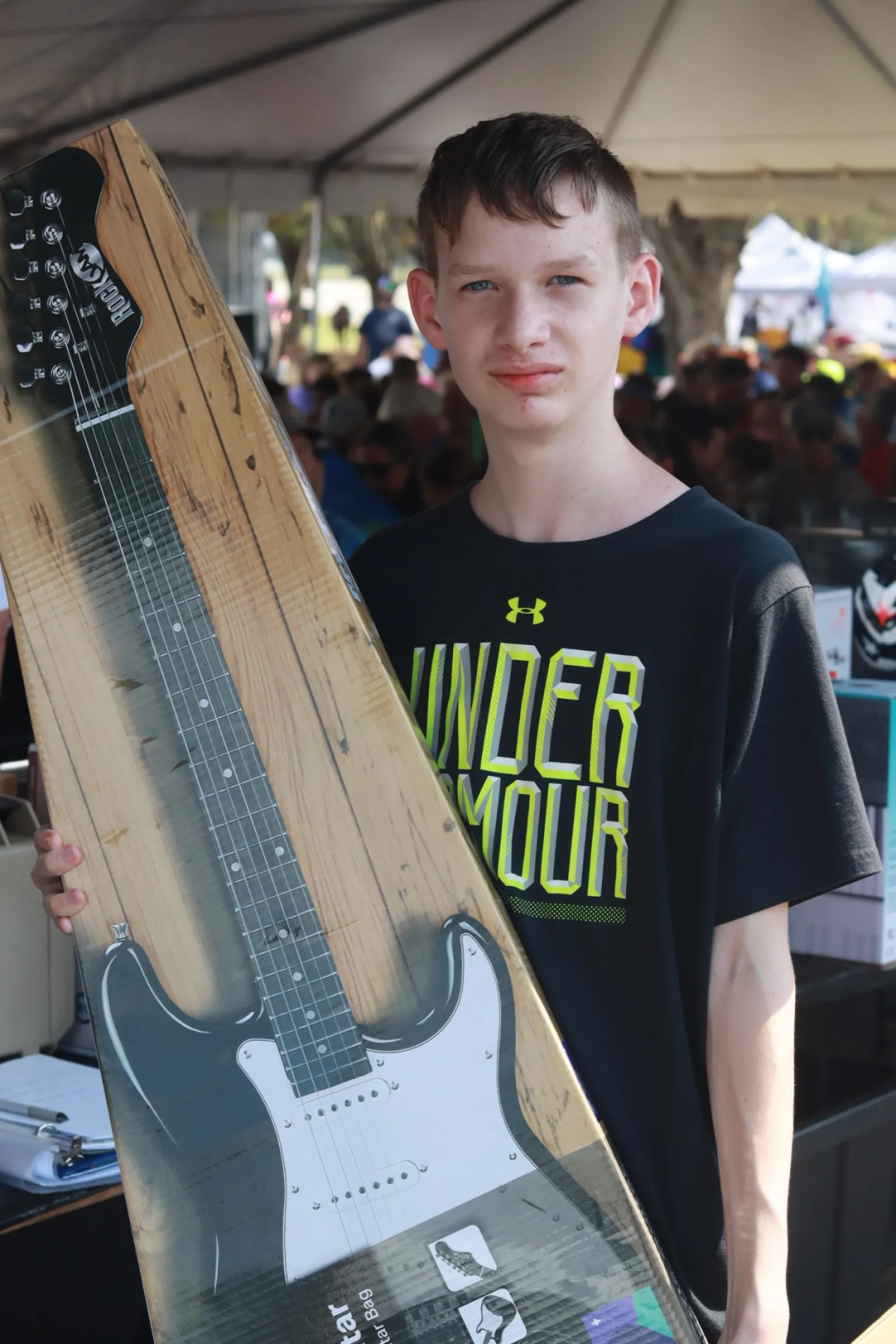 Boy holding boxed electric guitar at an outdoor event with tent and crowd in the background