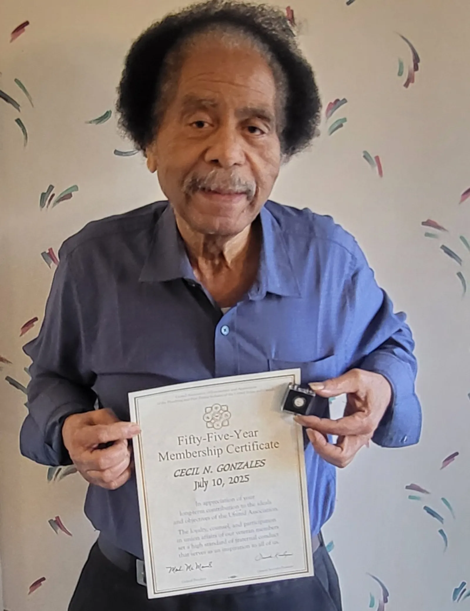 Elderly man holding a Fifty-Five-Year Membership Certificate and a pin for Cecil N. Gonzales, dated July 10, 2025.