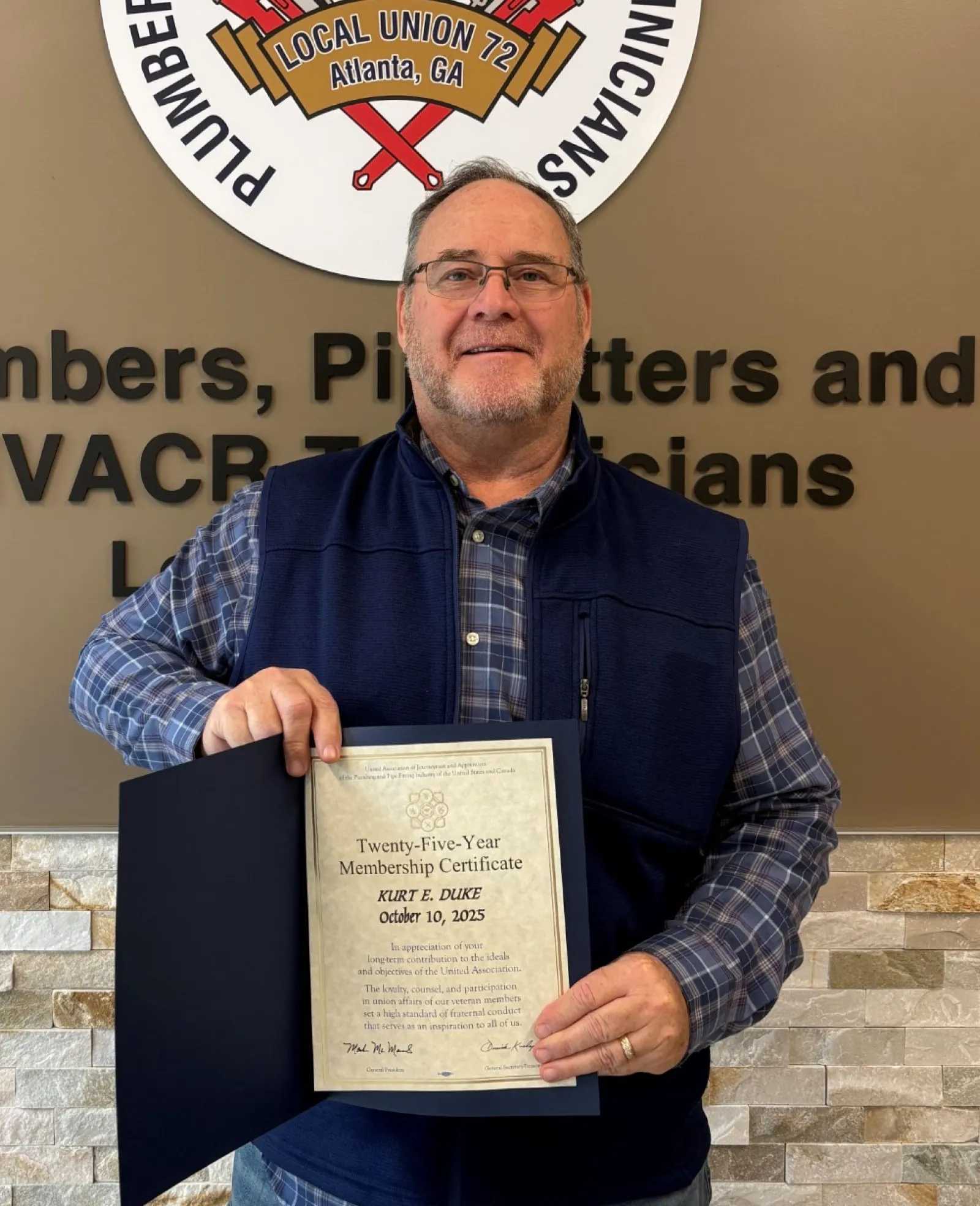 Man holding a certificate in front of a plumbers and pipefitters union sign and wall with text.