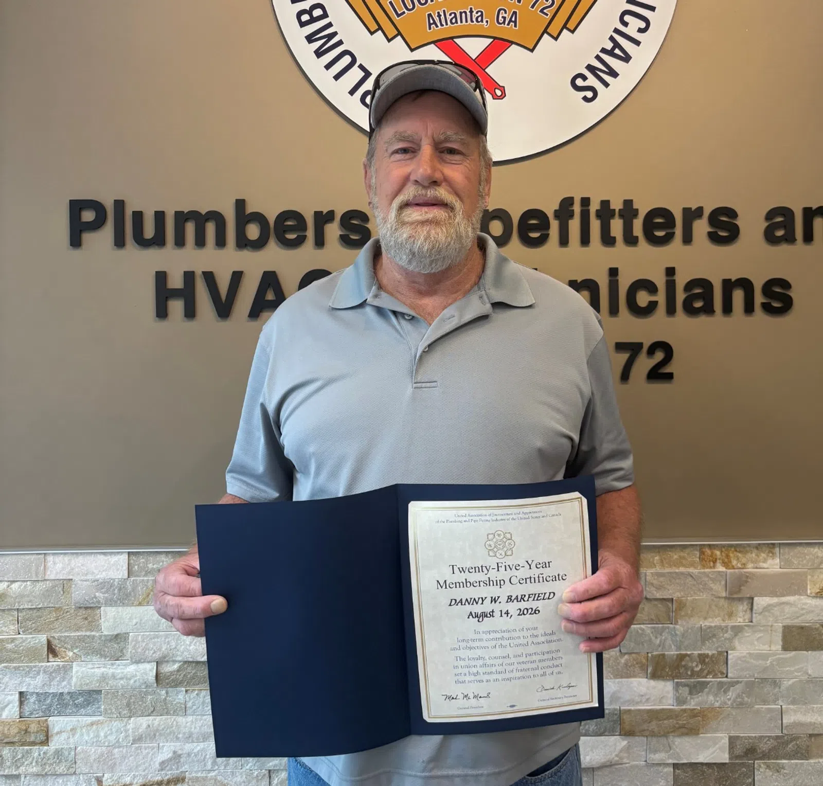 Man holding a certificate standing next to Plumbers and Pipefitters Local Union 72 sign on wall.