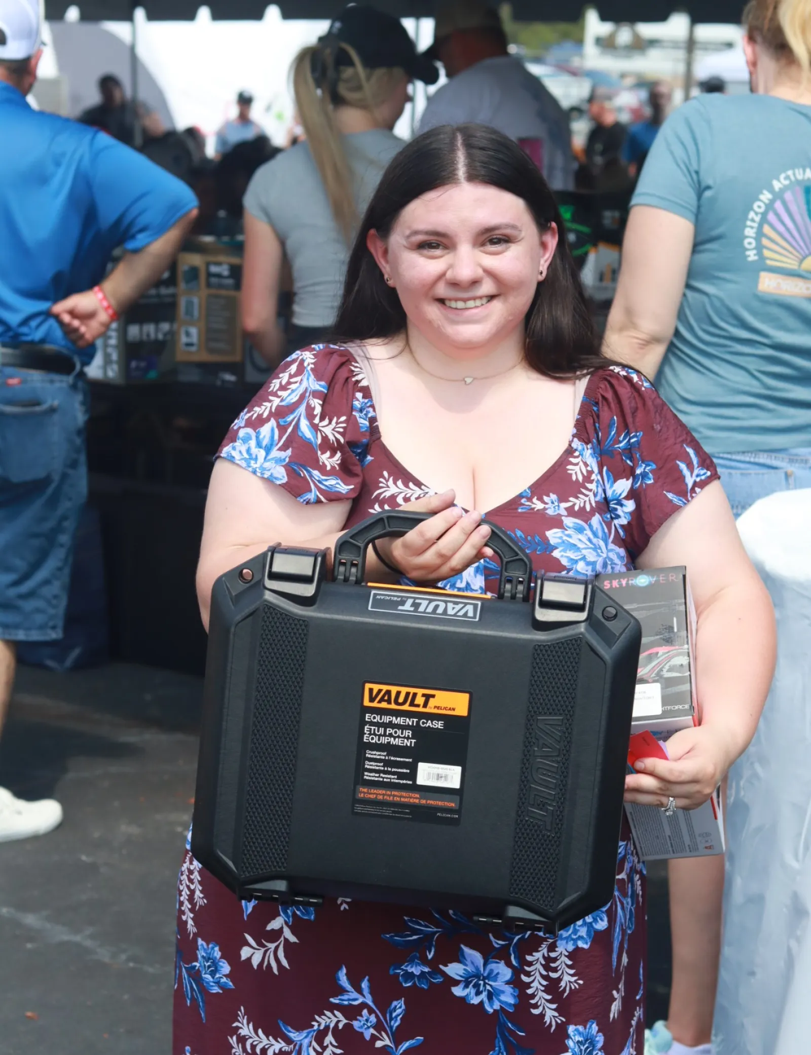Smiling woman in floral dress holding a black Vault equipment case at an outdoor event.