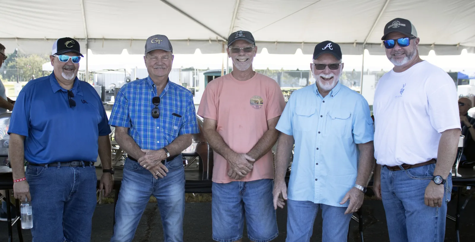 Five men wearing casual clothes and caps smiling under a white event tent on a sunny day.