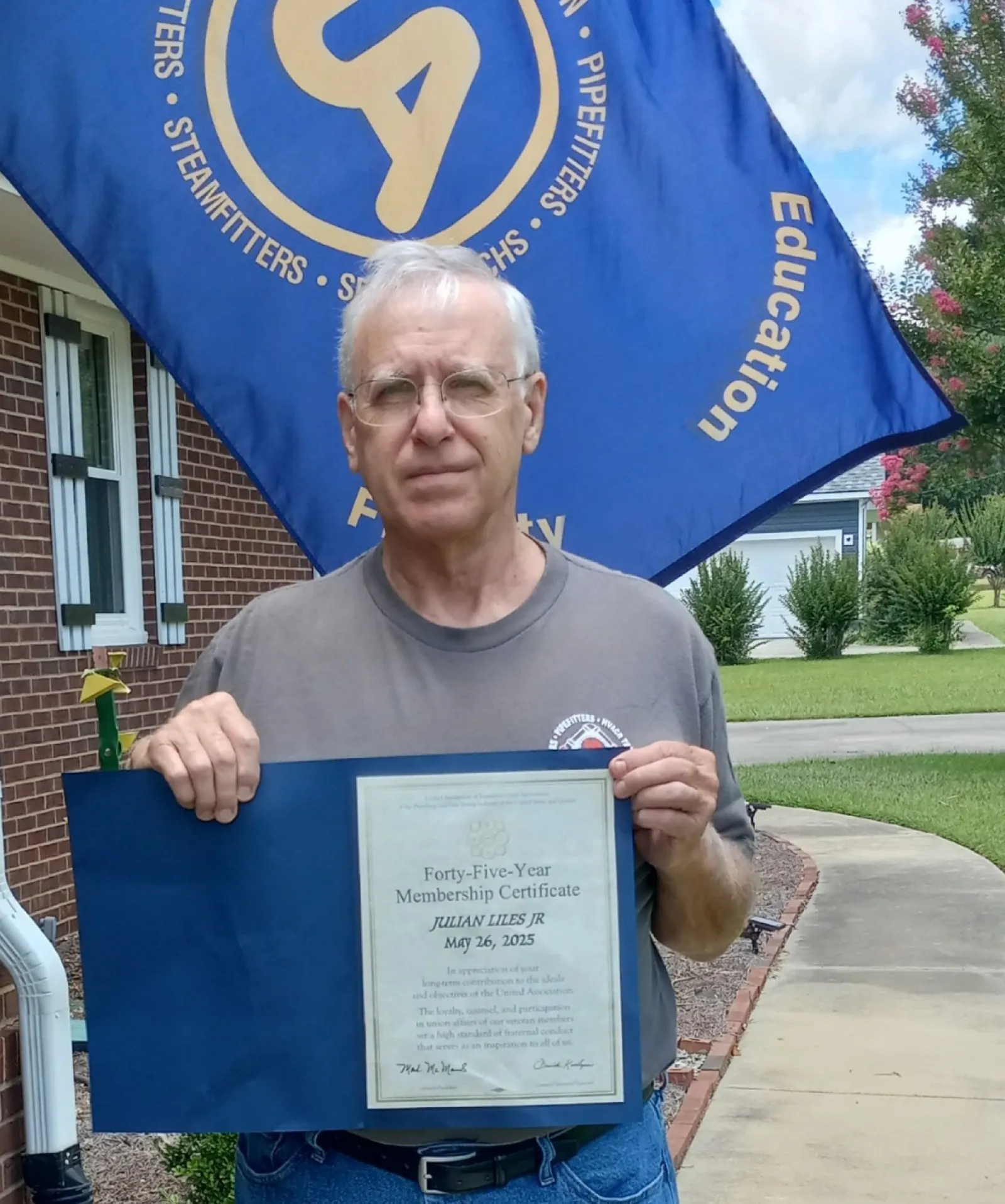 Man holding a Forty-Five-Year Membership Certificate outdoors with a blue flag in the background