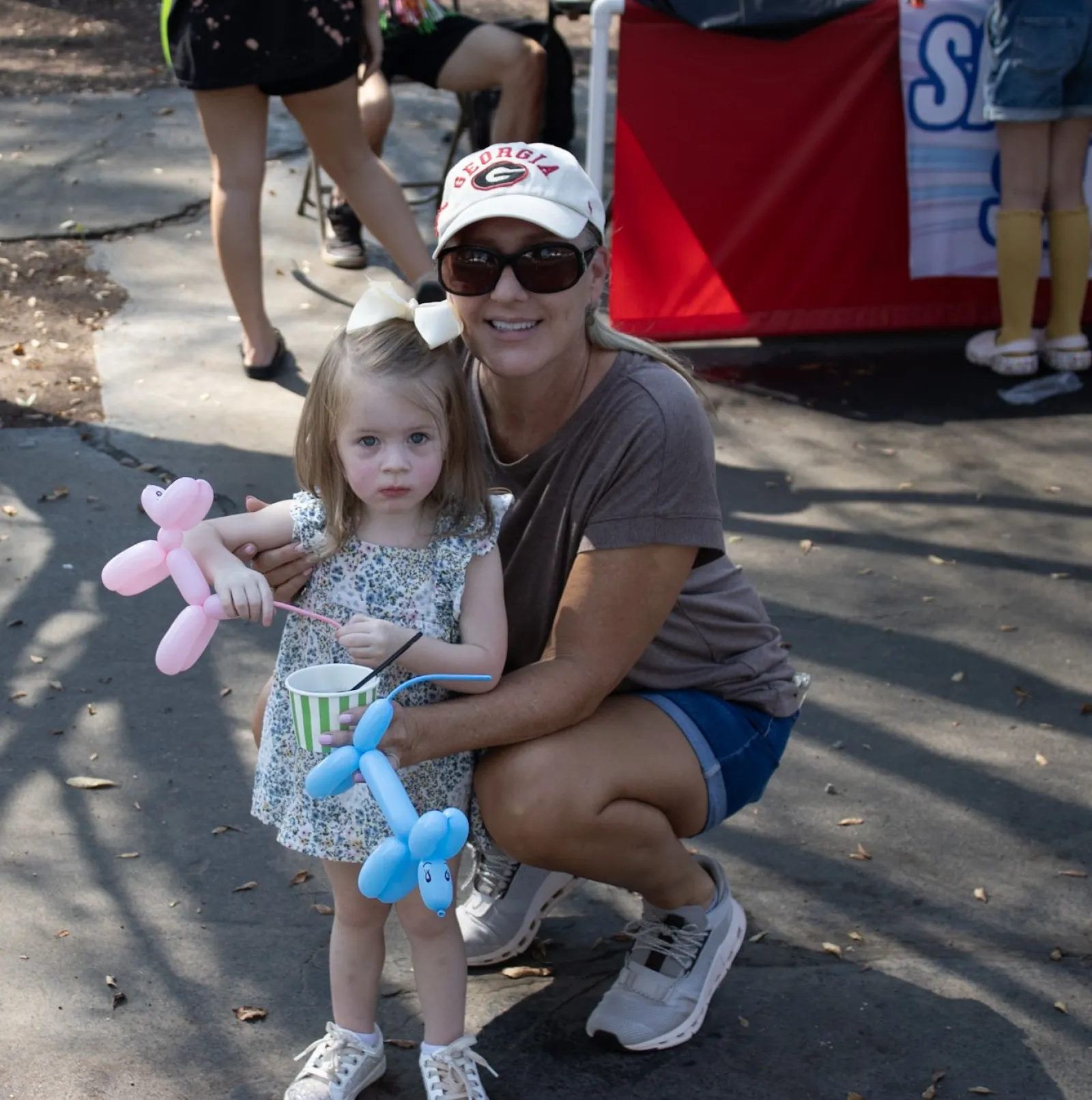 Young girl holding pink and blue balloon animals with woman in sunglasses and cap crouching beside her outdoors.