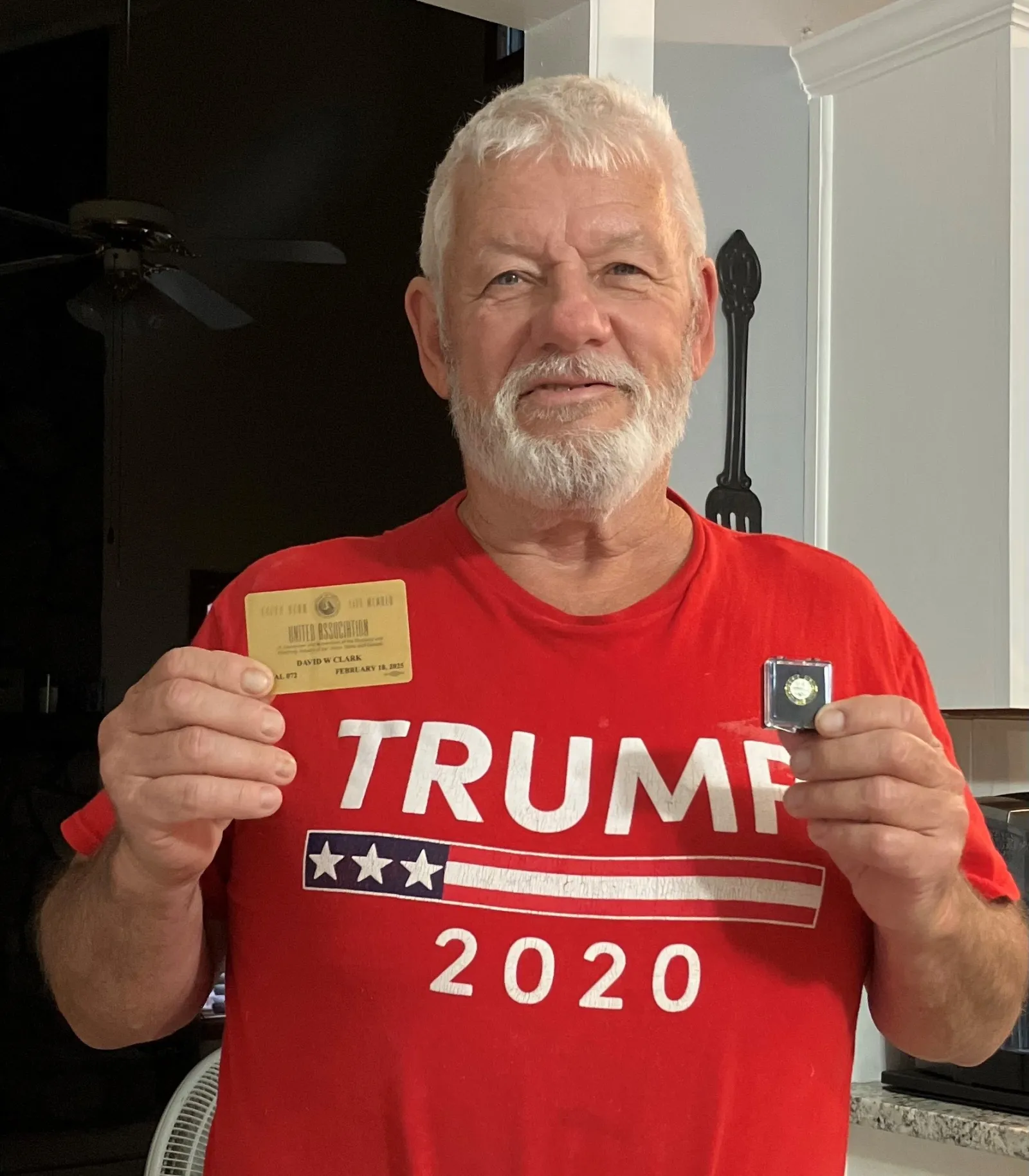 Older man with white beard wearing a red Trump 2020 shirt holding a card and a small medal in a kitchen.