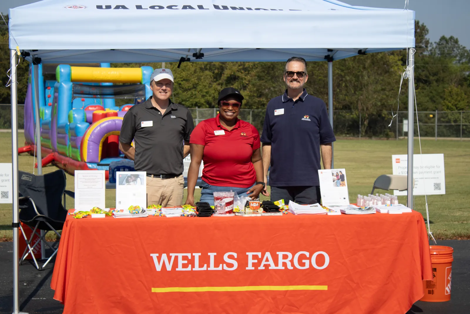 Three Wells Fargo representatives stand behind an orange table at an outdoor event under a white canopy tent.