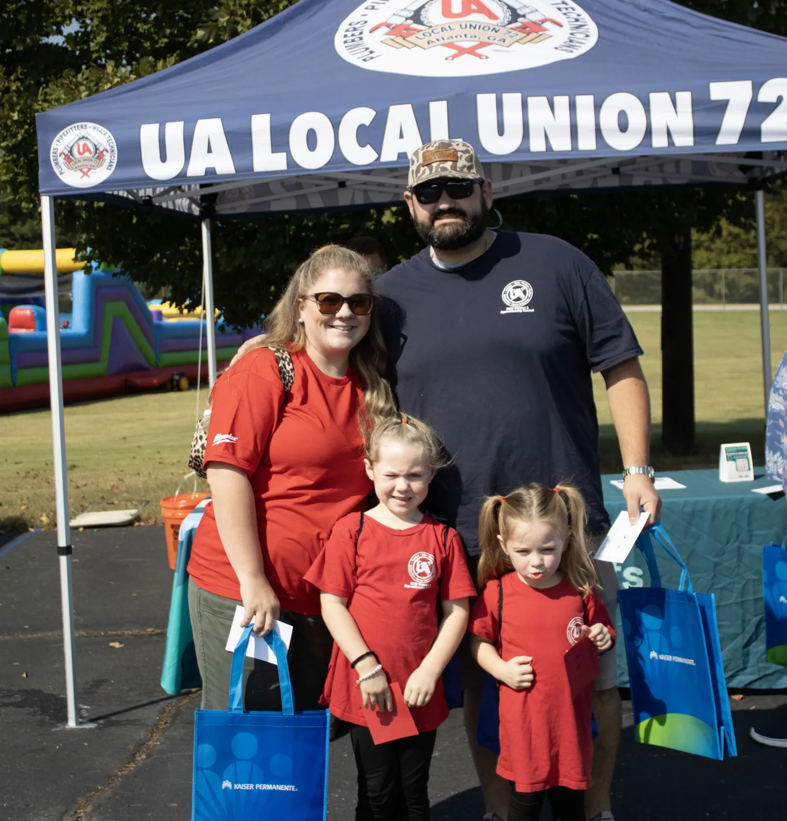 Family of four standing under UA Local Union 72 tent holding blue Kaiser Permanente bags outdoors.