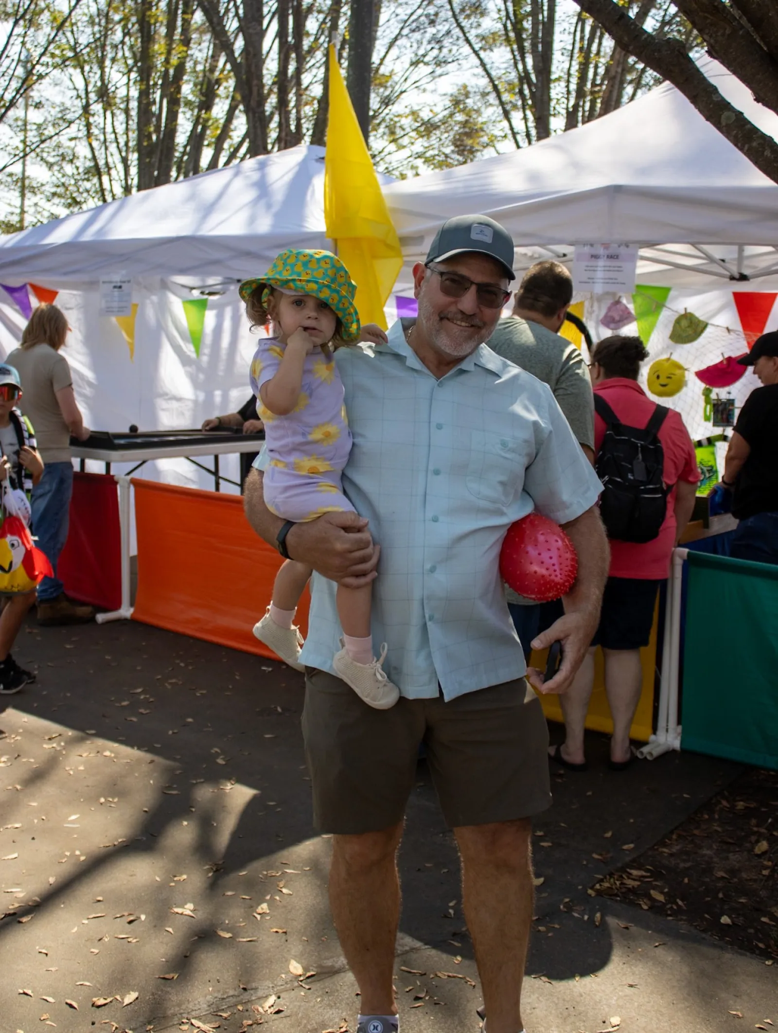 Smiling man holding a toddler in a colorful hat at an outdoor market with tents and festive decorations.